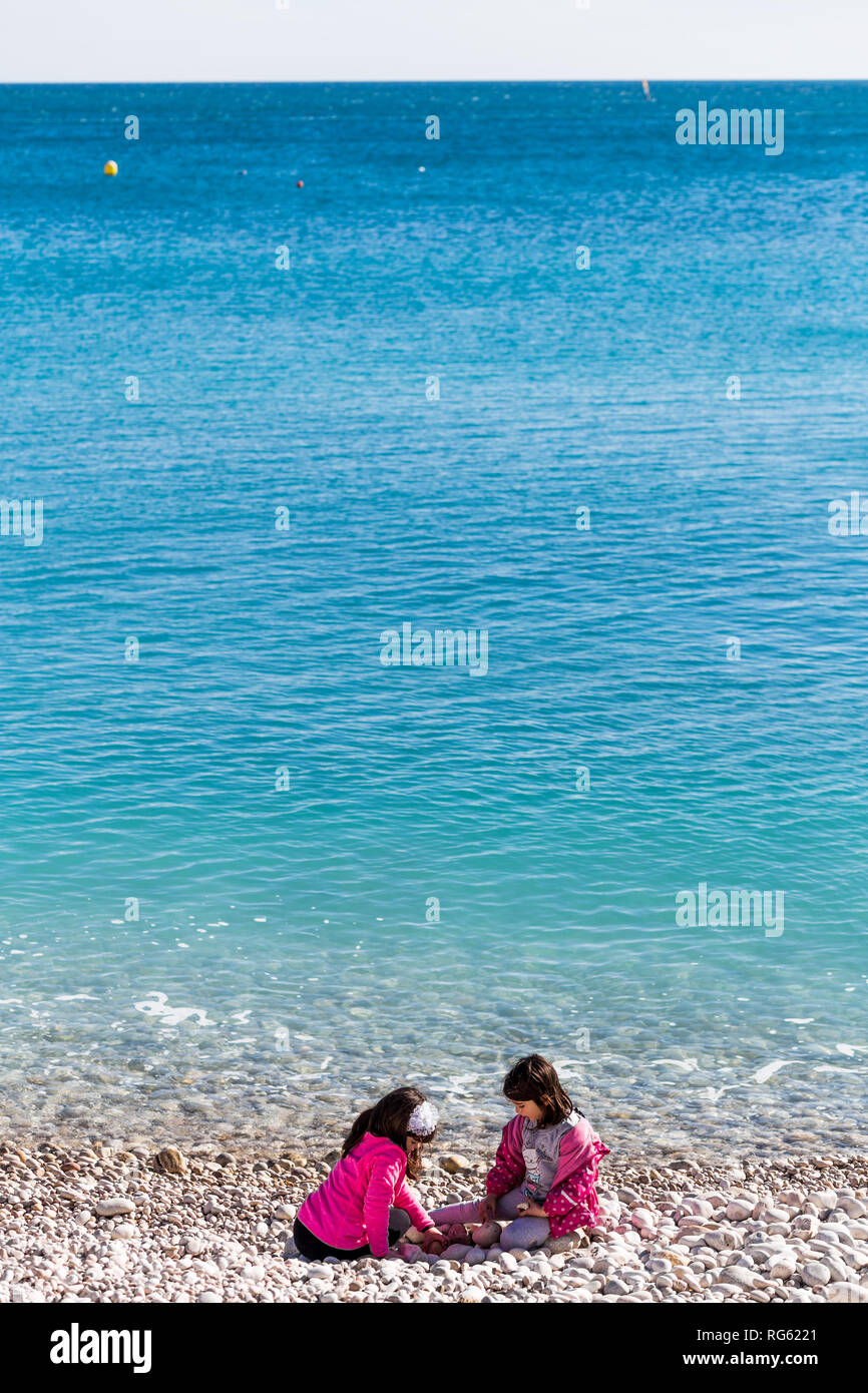 Two girls aged about 8 years old sitting on a pebble beach wearing pink jackets with blue sea in Spain. Stock Photo