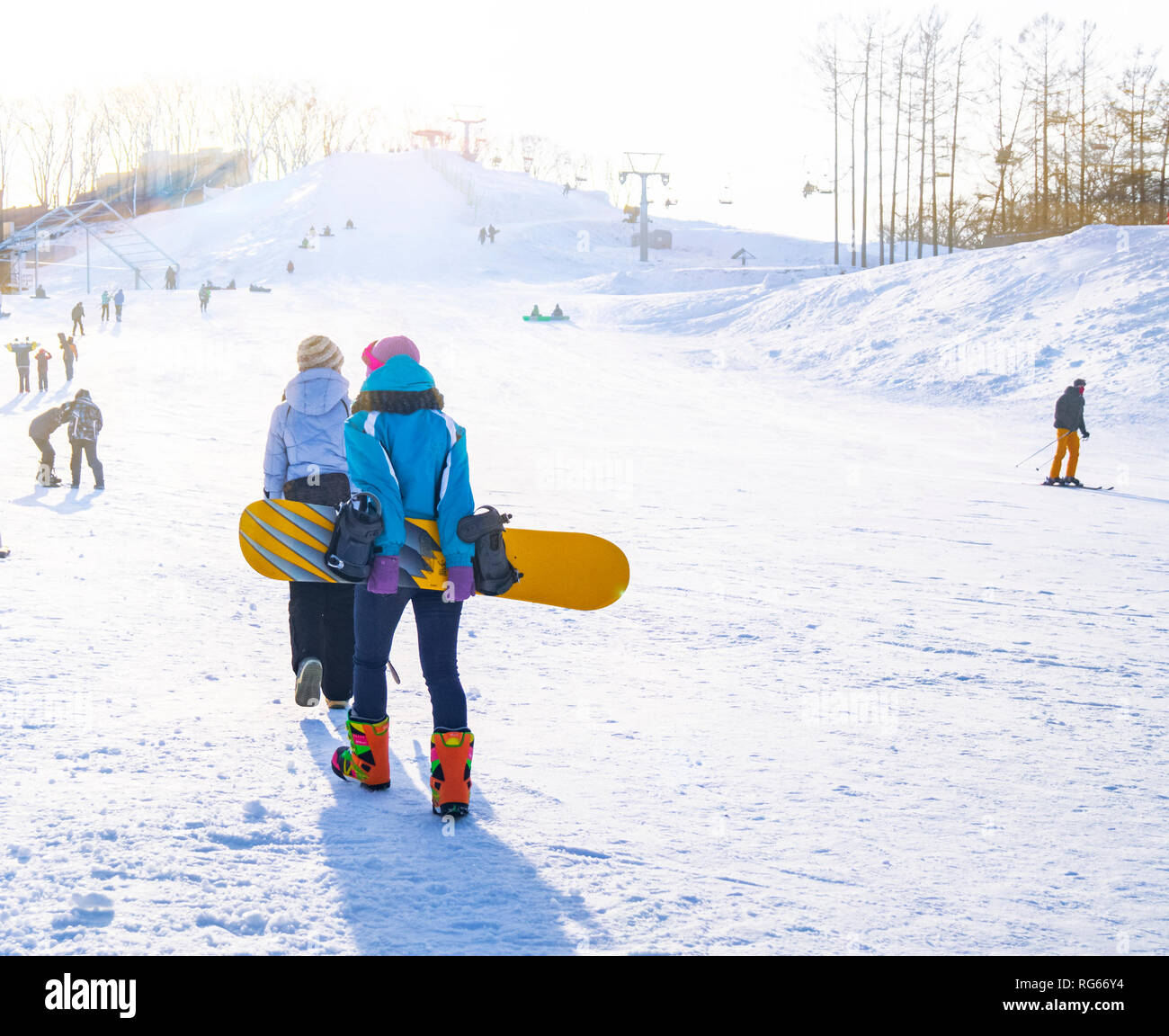 Young girls in special colored suits for winter fun and winter sports climb the hill to learn how to snowboard on a sunny day. Stock Photo