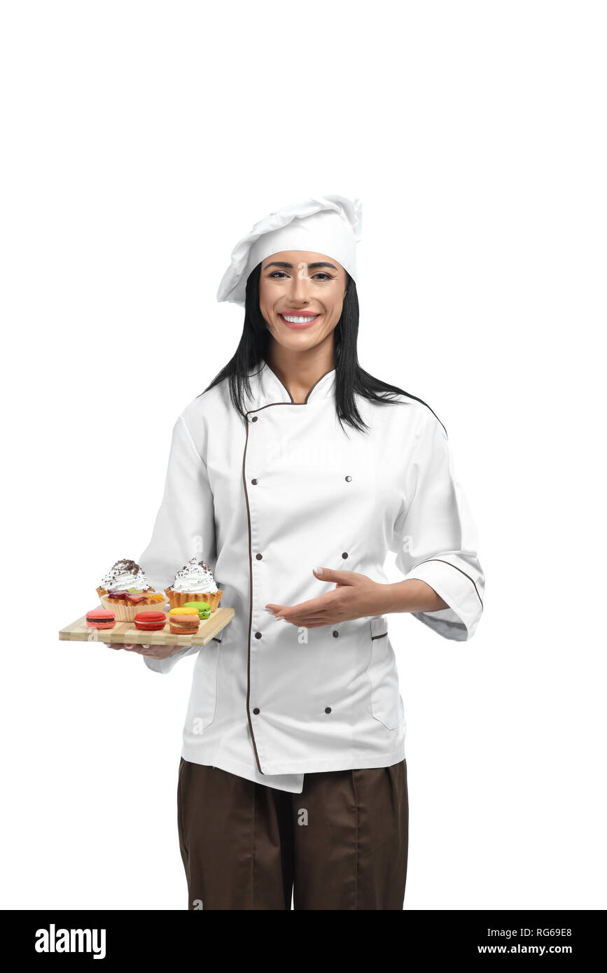 Happy woman holding tray with fantastic desserts and offering them to clients. Smiling pastry cook doing french sweets with whipped cream. Young confectioner in professional clothes liking her job. Stock Photo