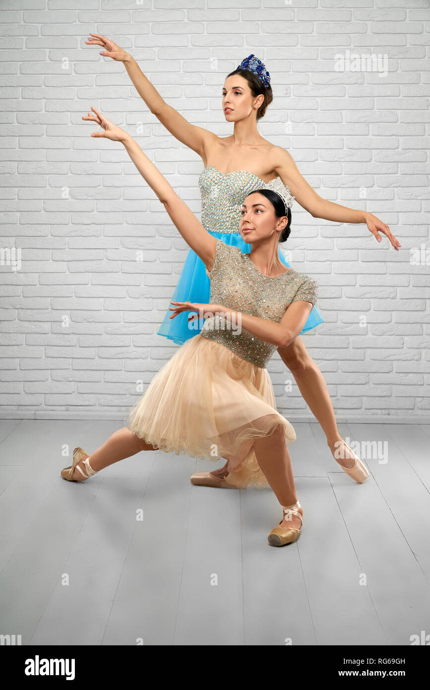 Front view of slim professional ballet dancers performing in studio. Young ballerinas in beige and blue dresses keeping hands up and looking above. Concept of ballet dance and elegance. Stock Photo