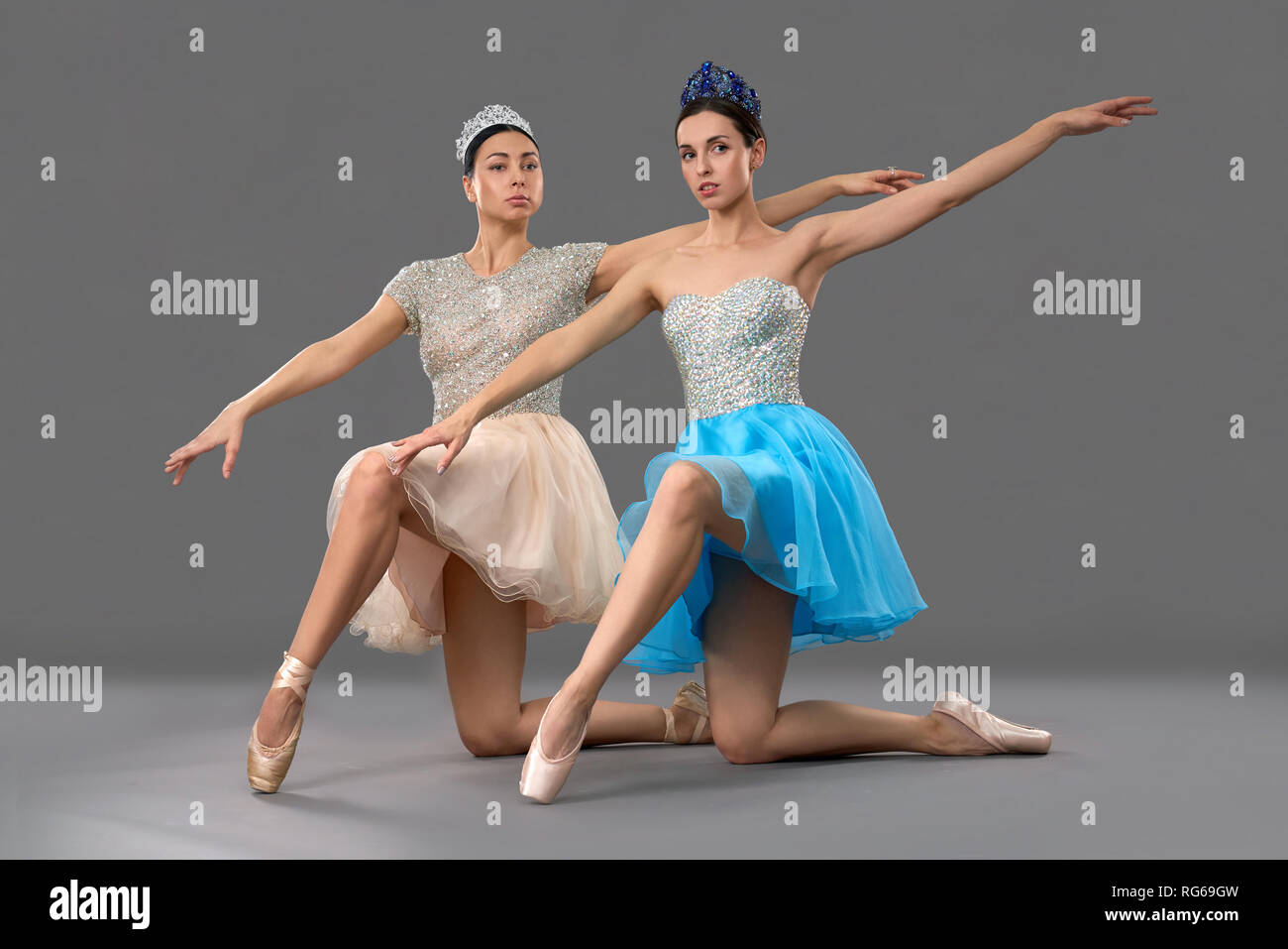 Adorable ballet dancers sitting on knees, raising one hand up and looking at camera in studio. Ballerinas in dresses and ballet shoes posing on grey background. Concept of performance and ballet. Stock Photo