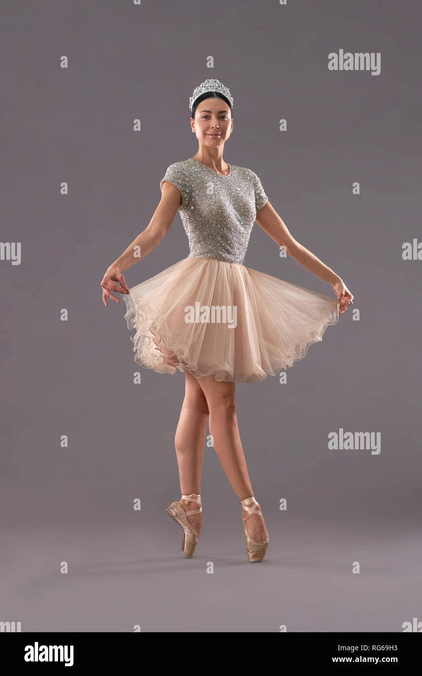 Front view of young ballerina in beige dress standing on socks, looking at  camera and smiling in studio. Ballet dancer keeping dress with fingers and  posing on grey background. Concept of ballet