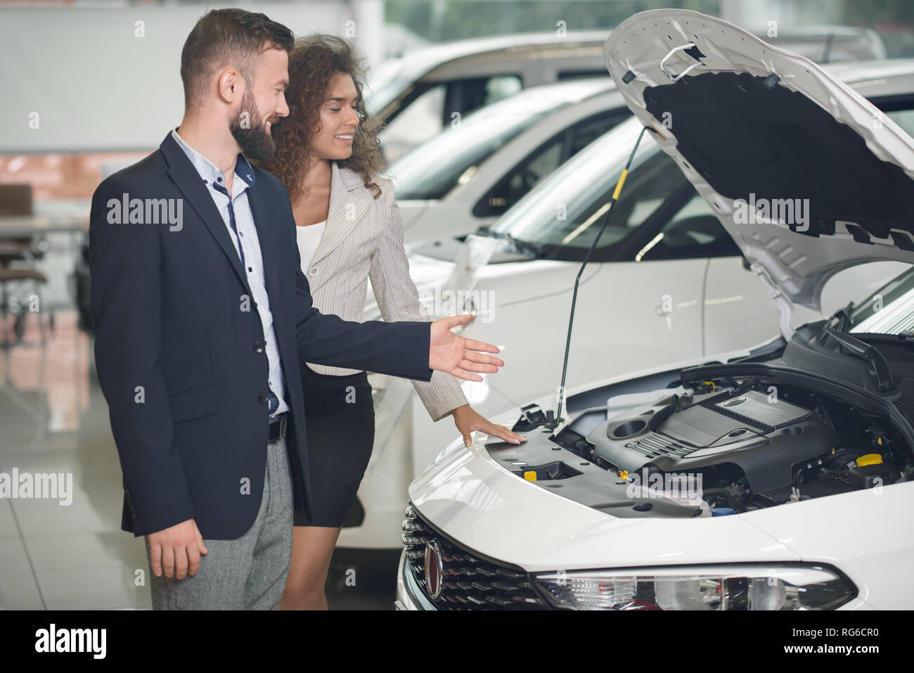 Car dealer showing to female client white automobile in auto showroom. Manager and beautiful woman observing together car under hood. Handsome bearded man wearing in dark blue jacket. Stock Photo