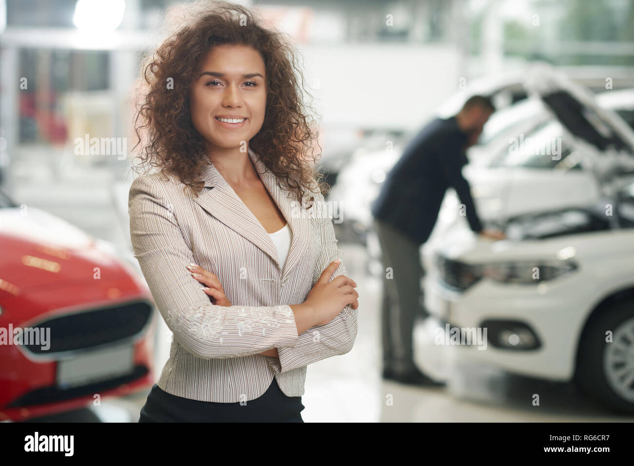 Beautiful woman standing in car showroom. Beautiful female manager of car dealership posing. Car dealer looking at camera, smiling. Unrecognizable man observing vehicle behind. Stock Photo