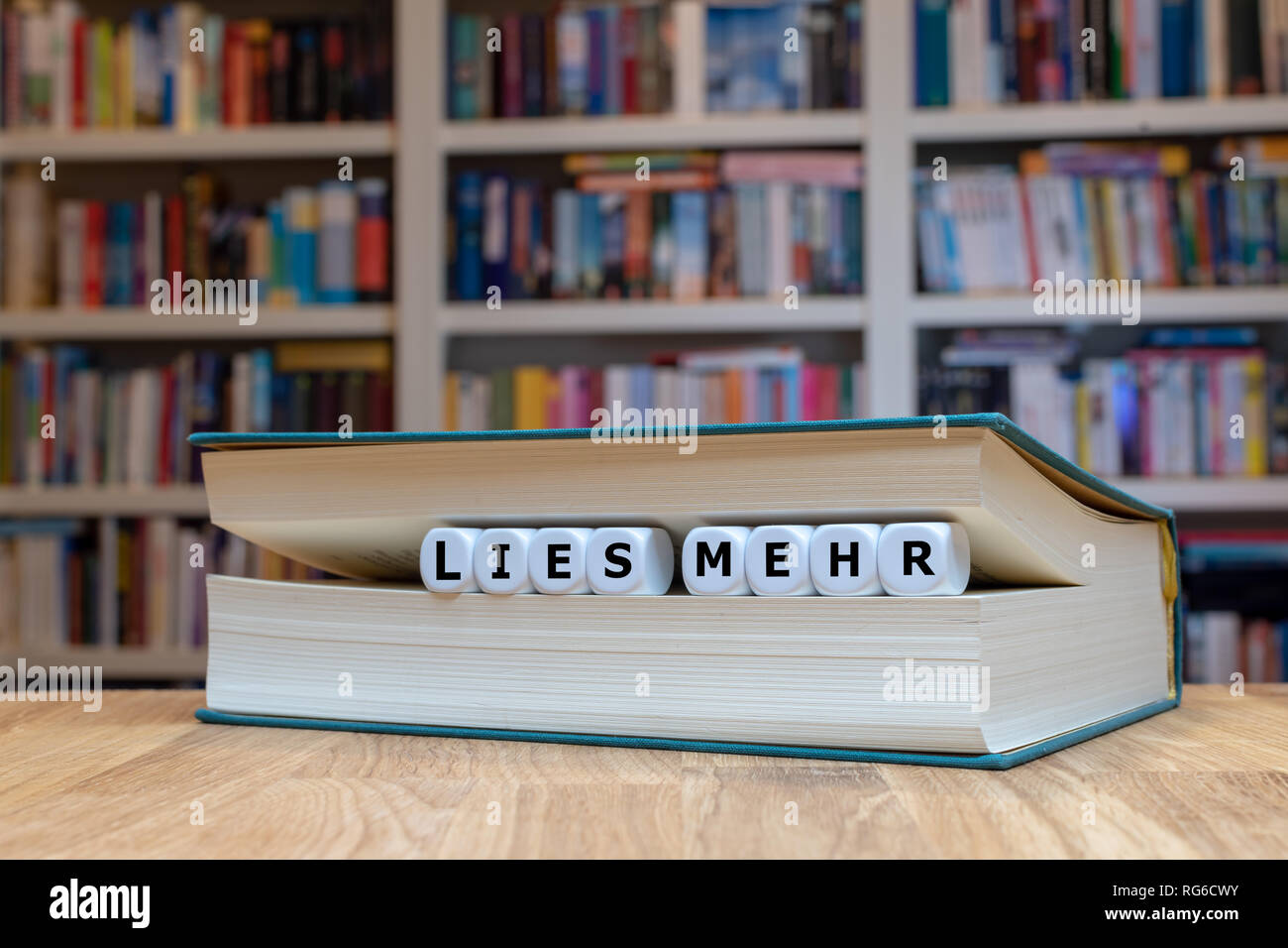 Würfel zwischen Buchseiten bilden die Worte 'lies mehr'. Das Buch liegt auf einer Holzoberfläche vor einem Bücherregal Stock Photo