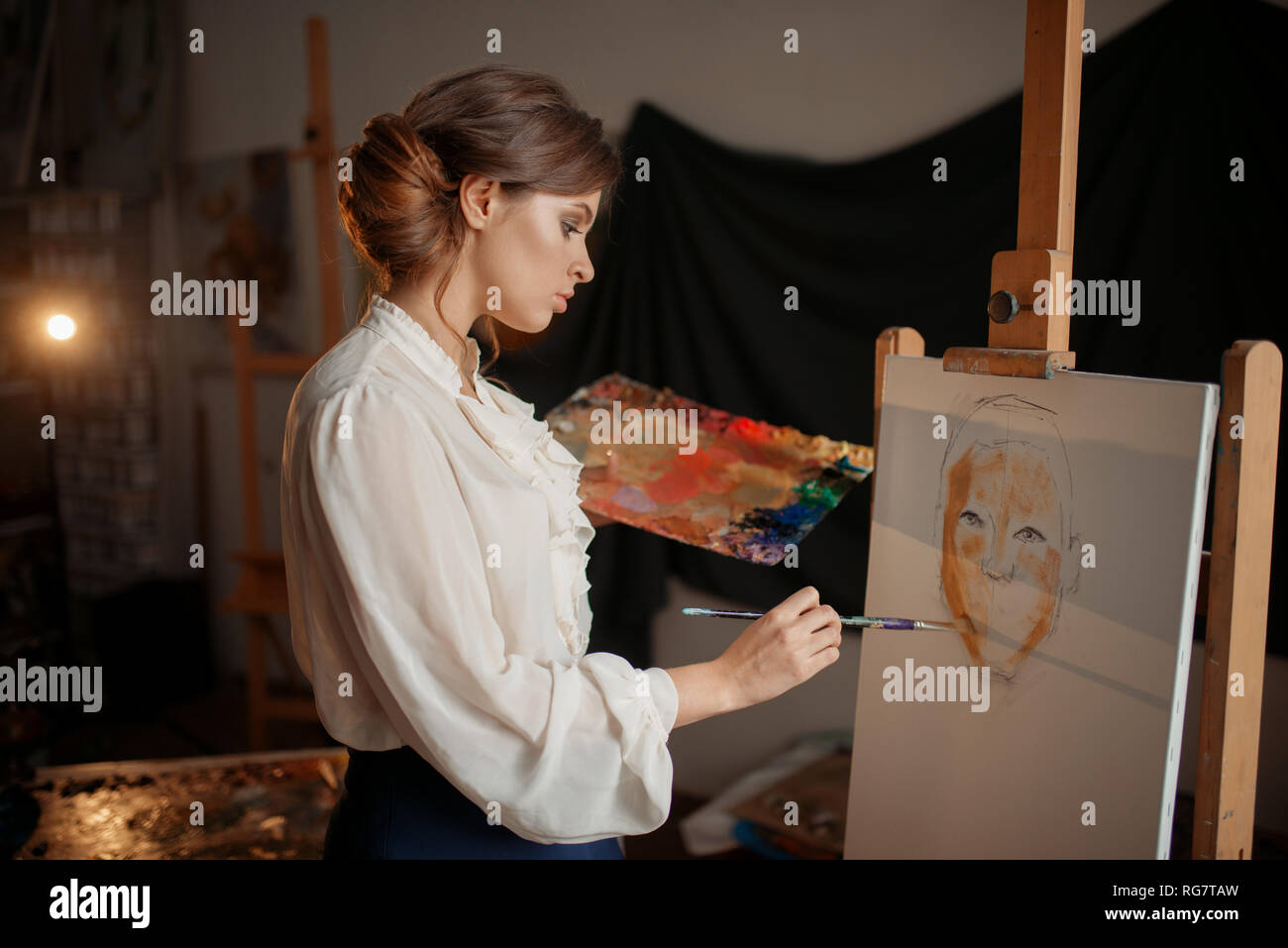 Female artist with color palette and brush standing against easel in ...