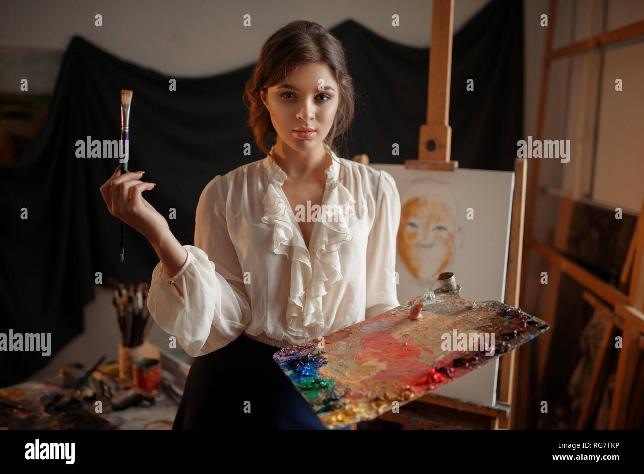 Female artist with color palette and brush standing against easel in ...