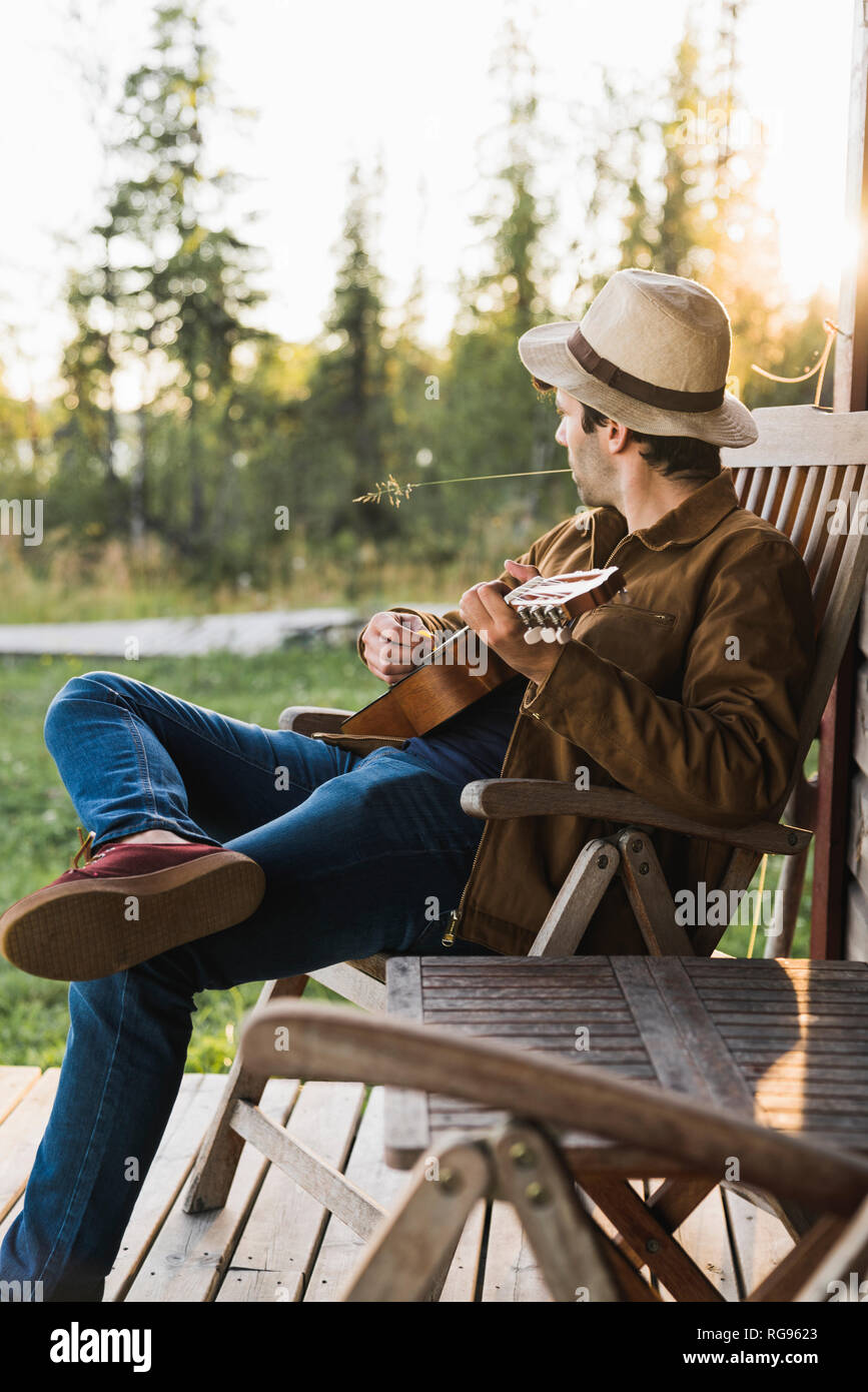 Young man sitting on veranda of a wood house, playing the ukulele Stock Photo