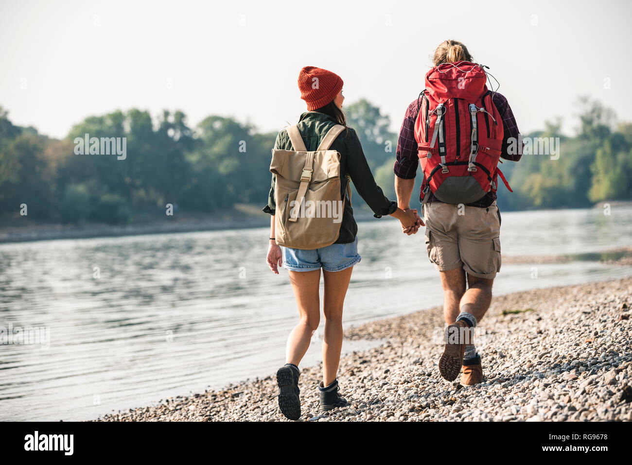 Rear view of young couple with backpacks walking hand in hand at the riverside Stock Photo
