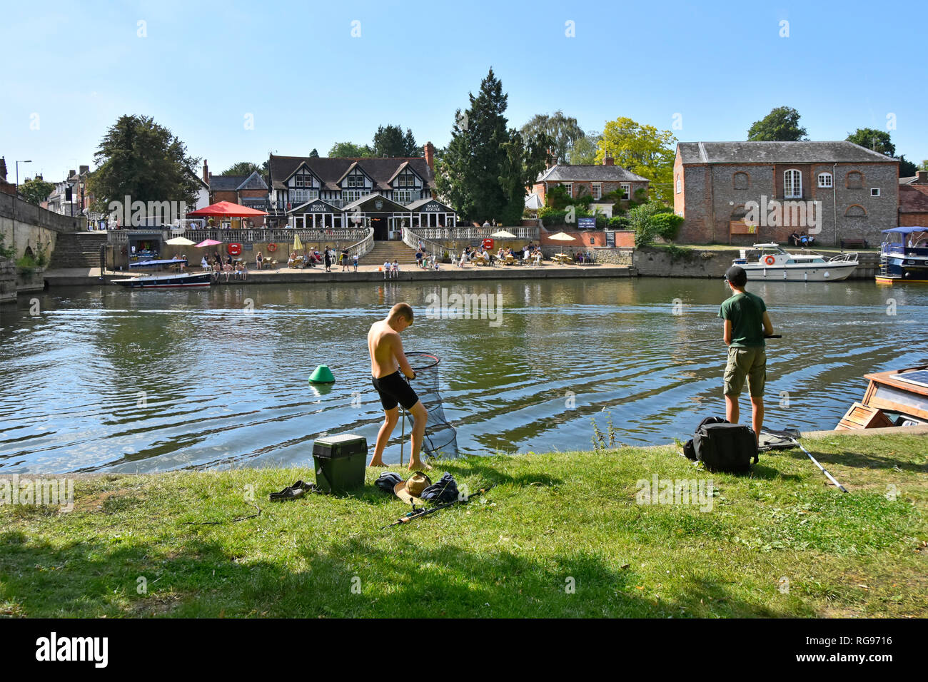 Two young teenagers boy fishing from shore River Thames on hot blue sky summer day with Boat House pub beyond at Wallingford Oxfordshire England UK Stock Photo