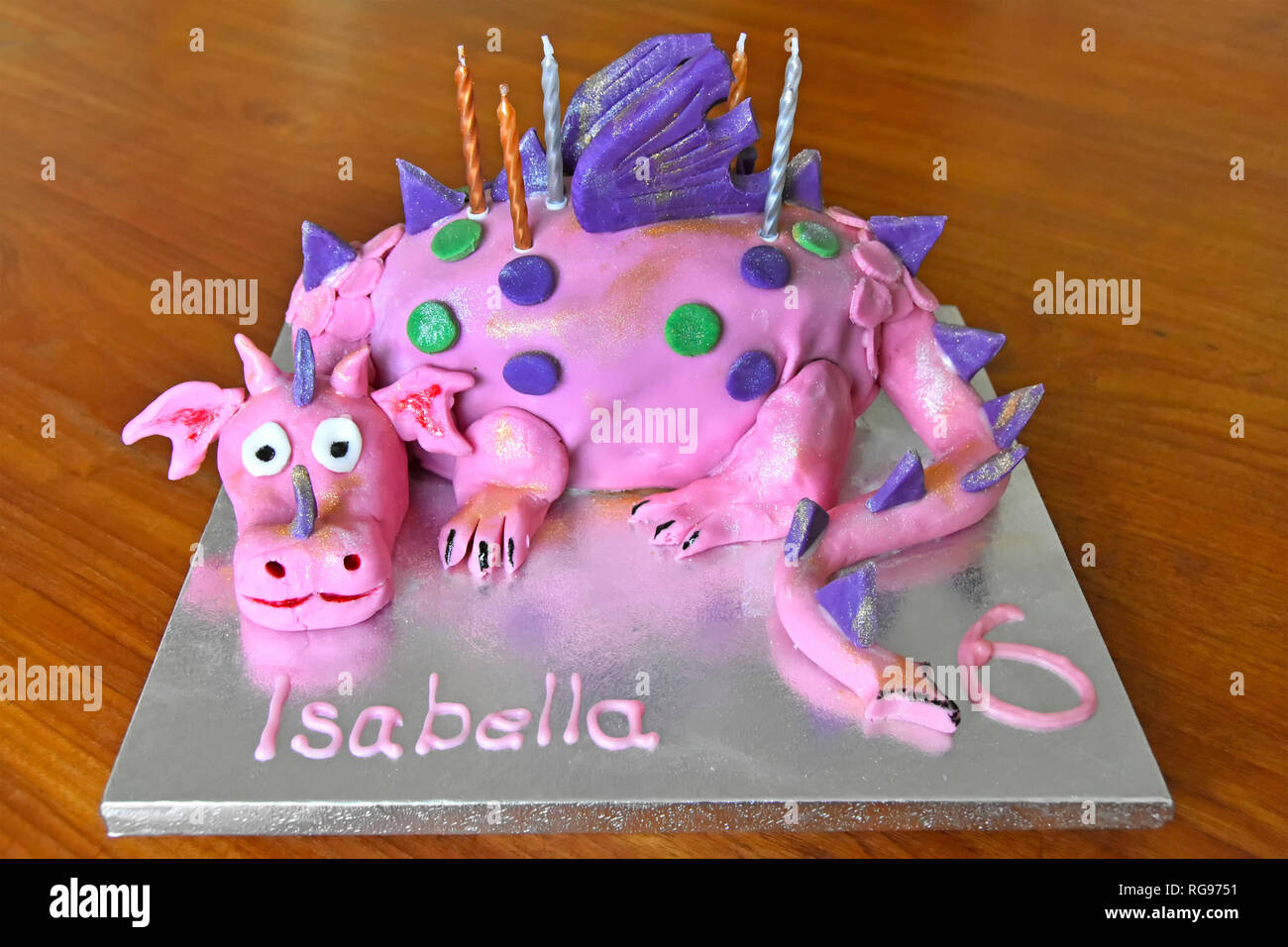 Home made cake for six years old childs birthday celebration with candles on cake board with table background featuring caricature of pink dragon Stock Photo
