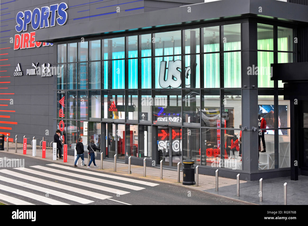 Dusk view of customers at Sports Direct store building entrance & glass shop front bollards protect from ramming retail park Thurrock Essex England UK Stock Photo