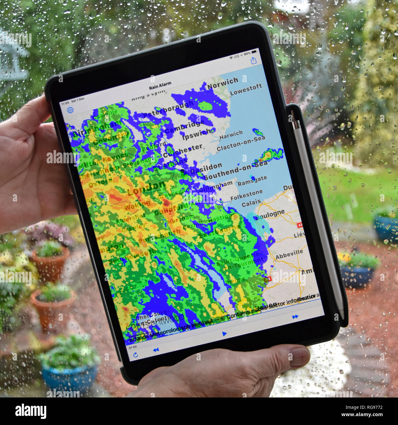Woman hands holding ipad pro tablet rain alarm app uses real time colour radar data superimposing rainfall on local map wet weather on window Essex UK Stock Photo