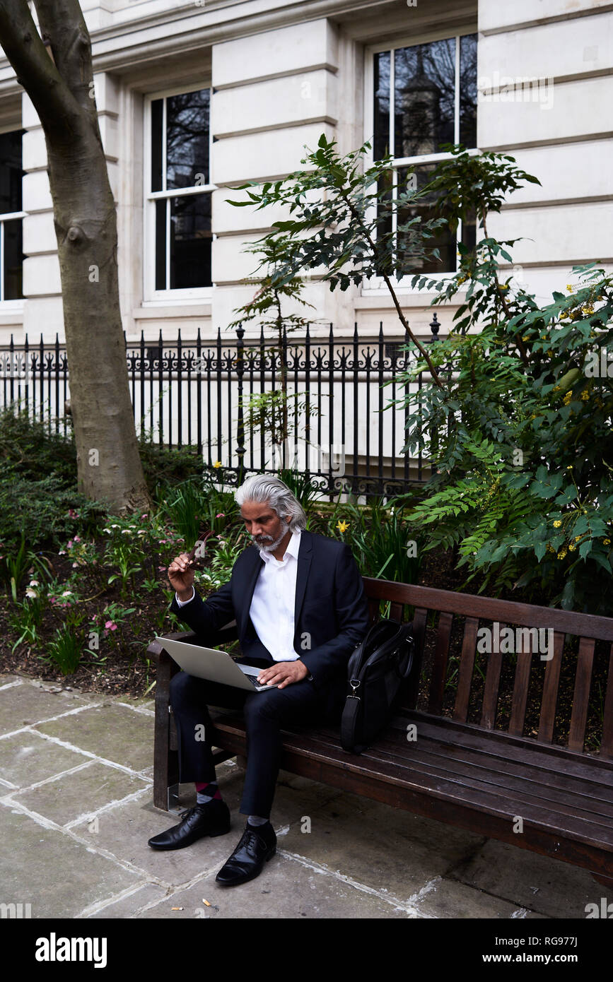 Senior businessman sitting on bench outdoors working on laptop Stock Photo