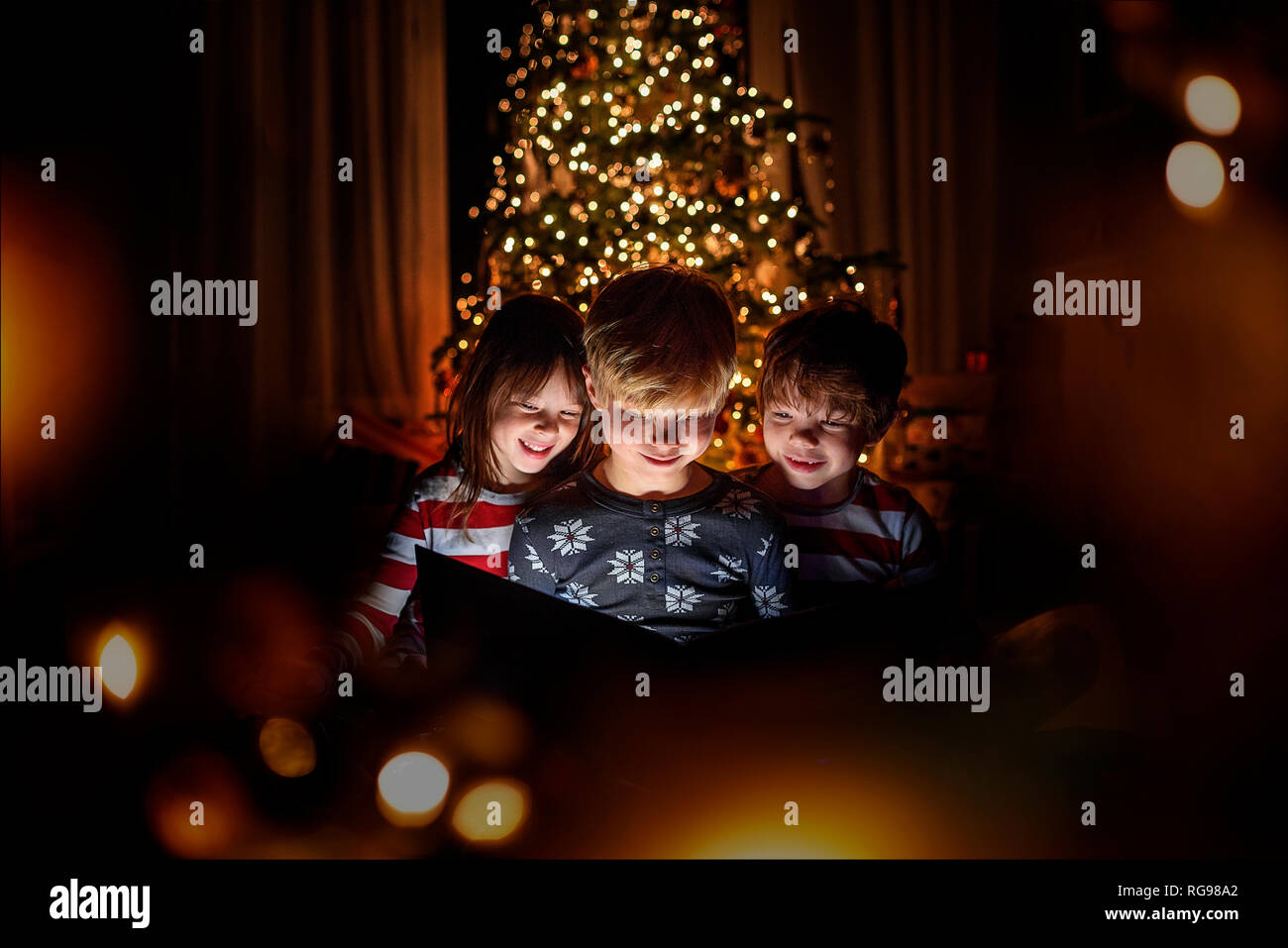 Three children sitting in front of a Christmas tree reading a book Stock Photo