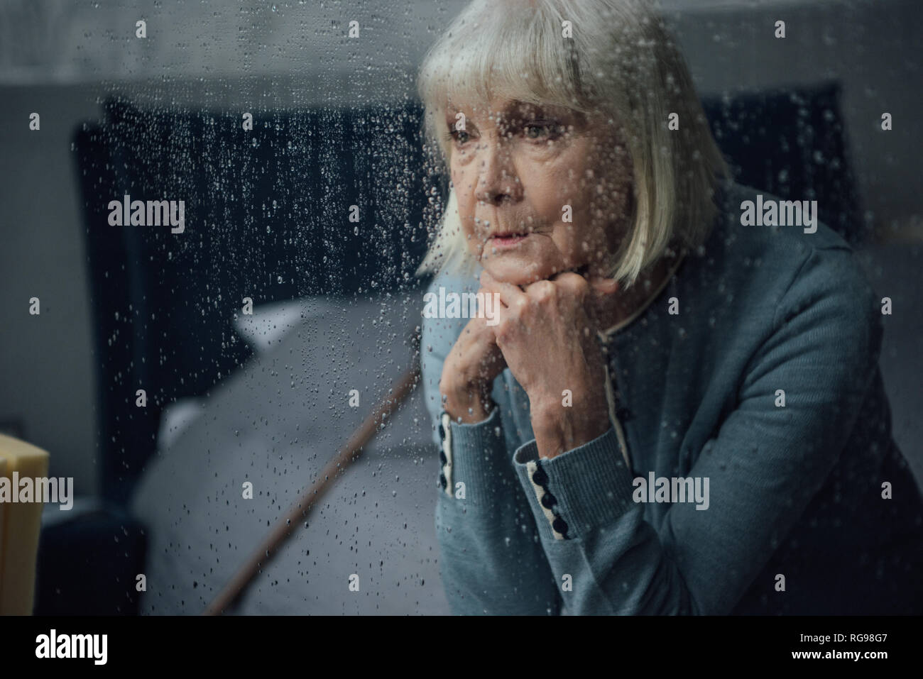 upset senior woman sitting and propping chin with hands at home through window with raindrops Stock Photo