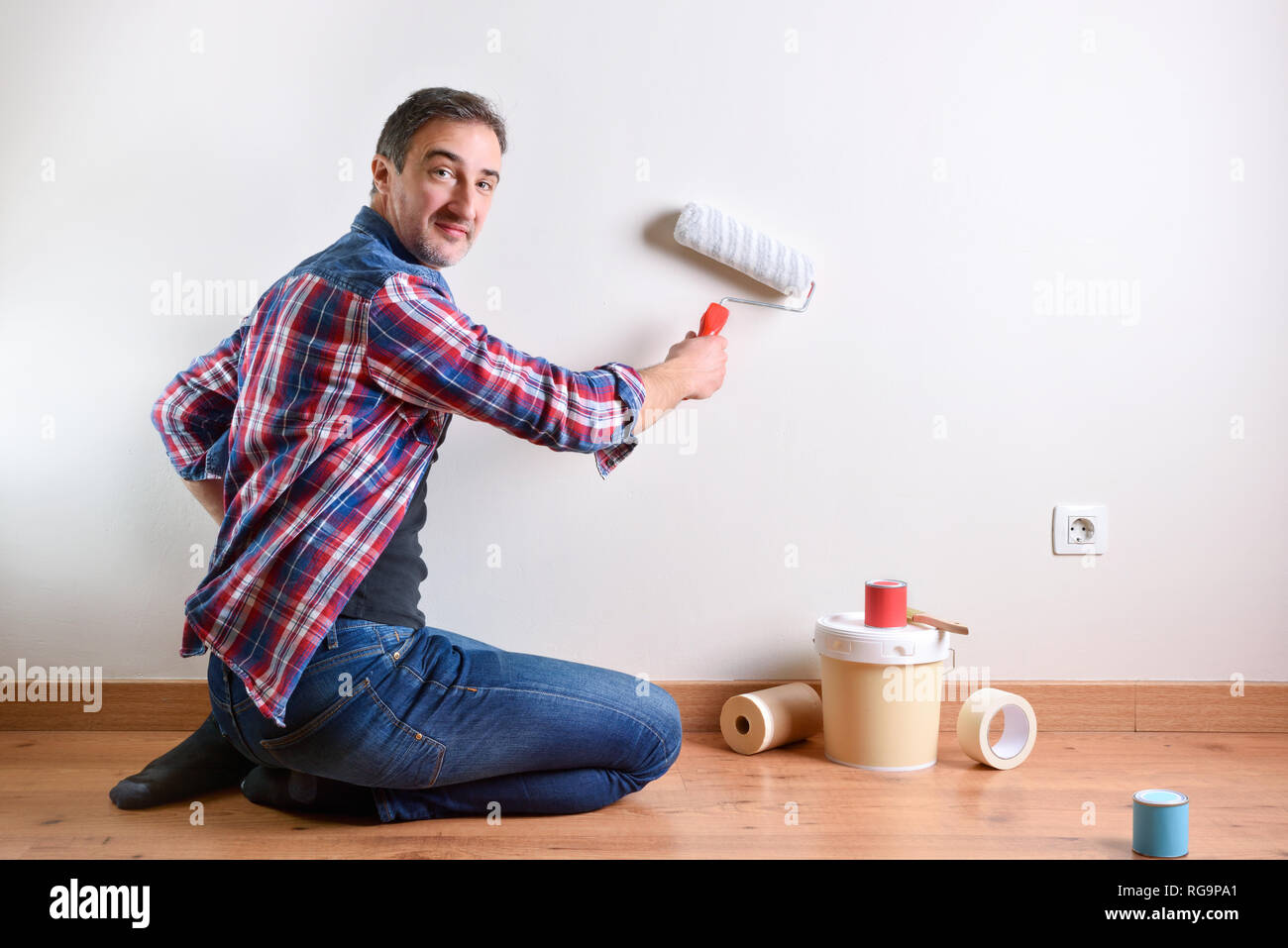 Smiling man kneeling on a parquet floor with paint material painting his house. Front view. Horizontal composition. Stock Photo
