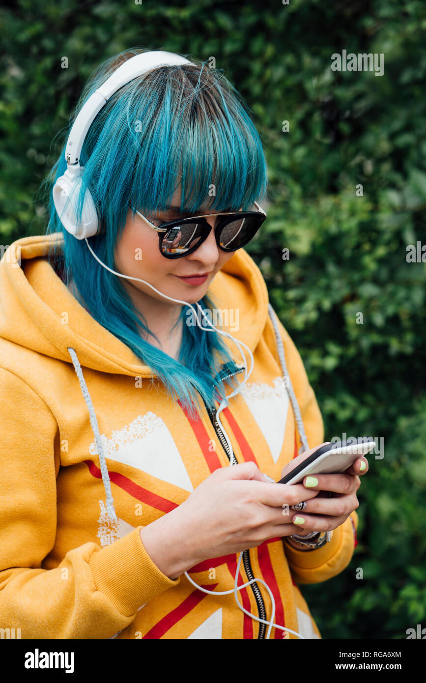 Portrait of young woman with dyed blue hair listening music with headphones looking at smartphone Stock Photo