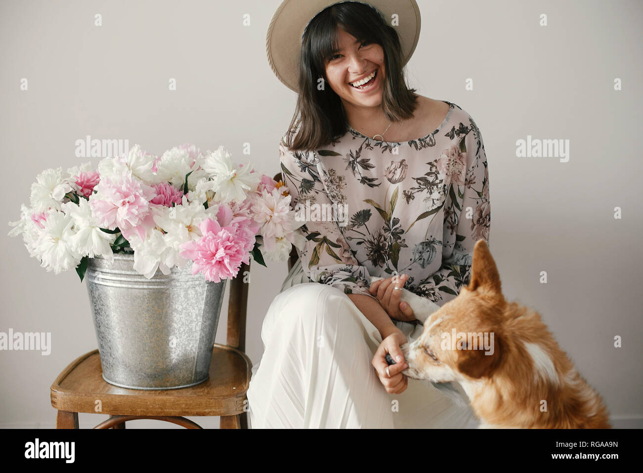 Stylish boho girl sitting at metal bucket with peonies on rustic wooden chair and playing with cute golden dog.Beautiful hipster woman playing with he Stock Photo
