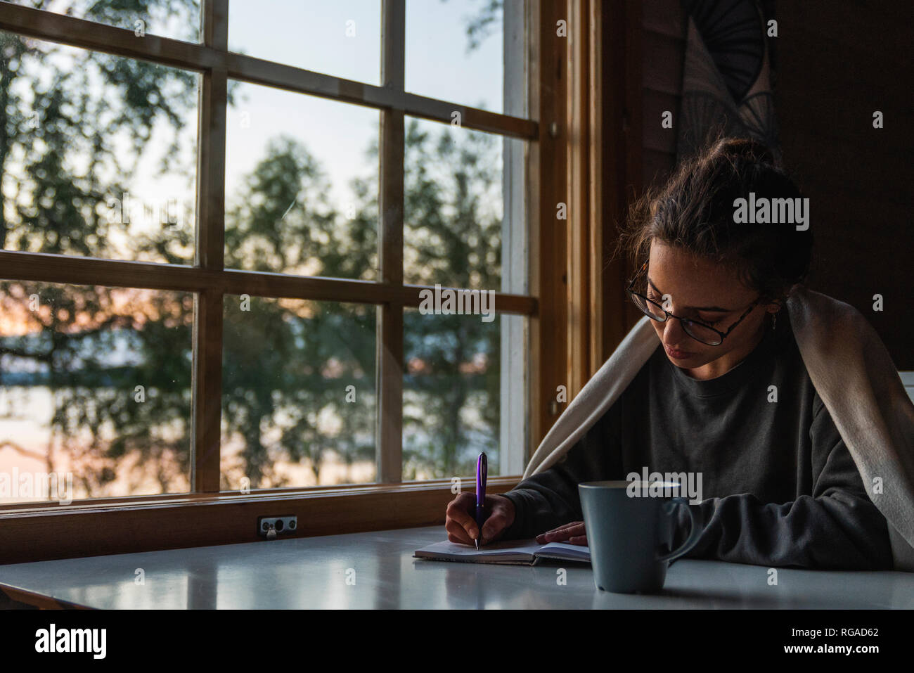 Finland, Lapland, young woman sitting at the window at a lake writing into diary Stock Photo