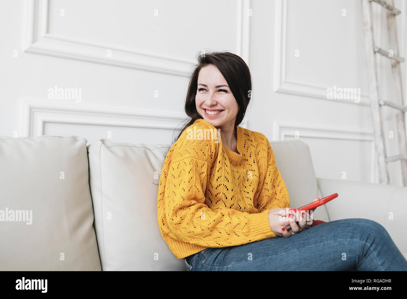 Portrait of enjoyed young woman with smartphone sitting on the couch at new home looking at distance Stock Photo