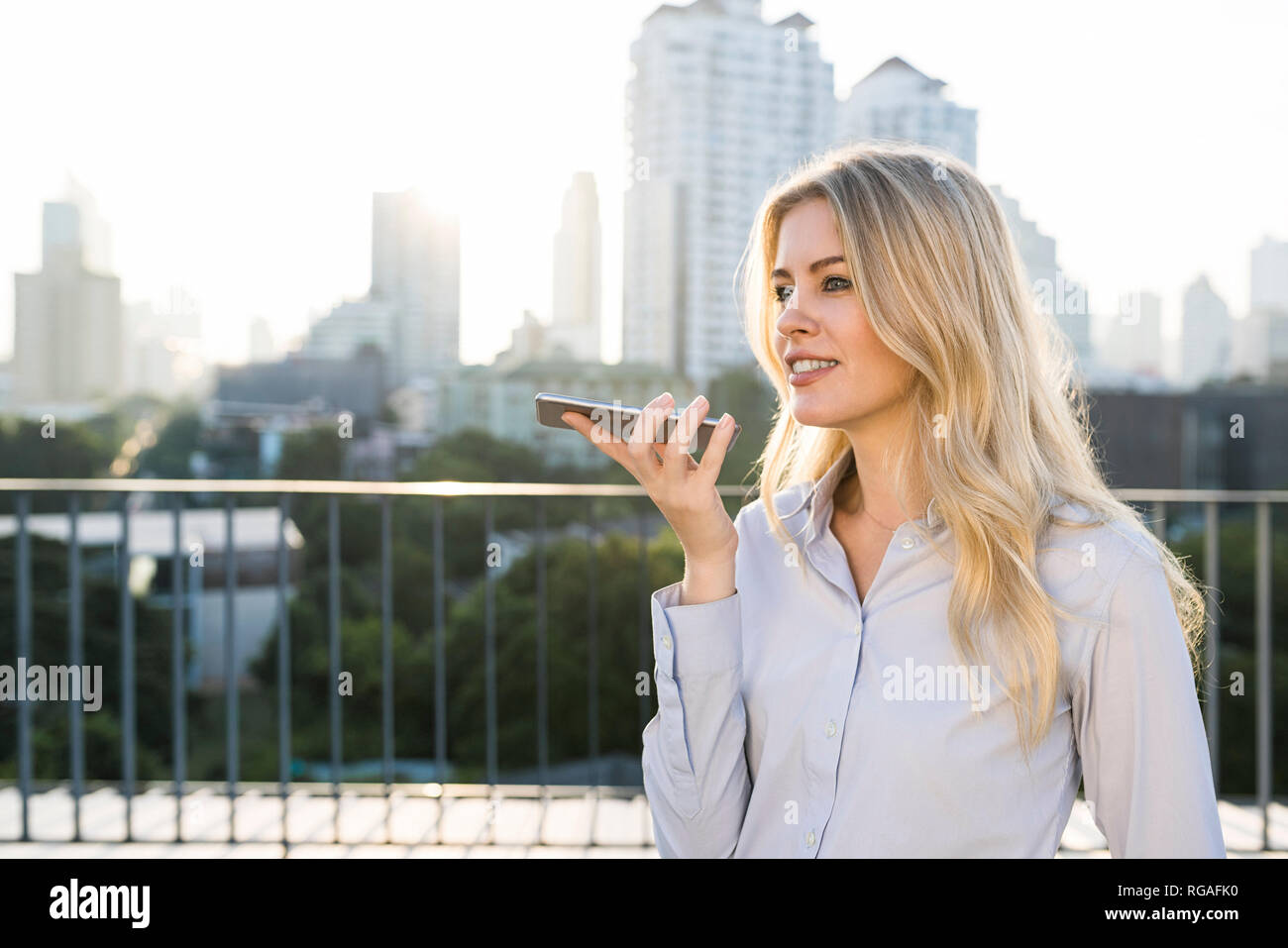 Blonde business woman speaking into smartphone on city rooftop Stock Photo