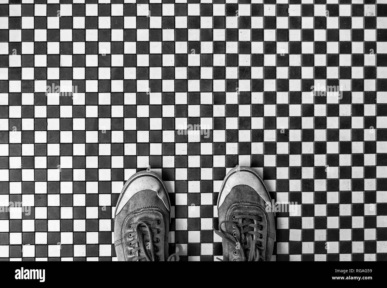 Looking down from above onto a pair of worn sneakers or shoes standing on a black and white checkered mosaic tiled floor with copy space Stock Photo