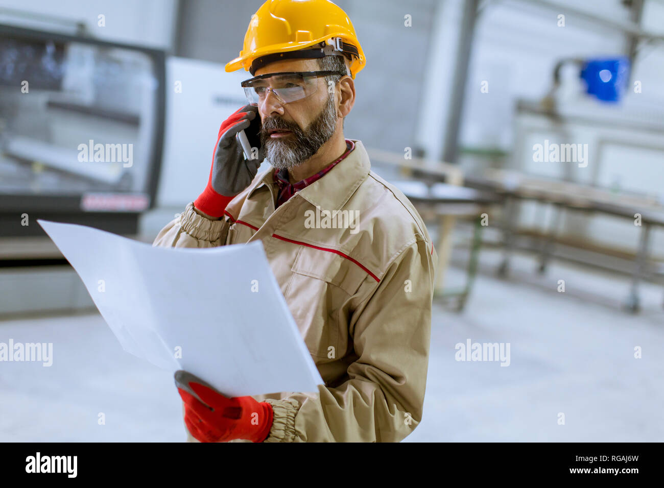 Handsome mature engineer looking at plan and using mobile phone in the factory Stock Photo