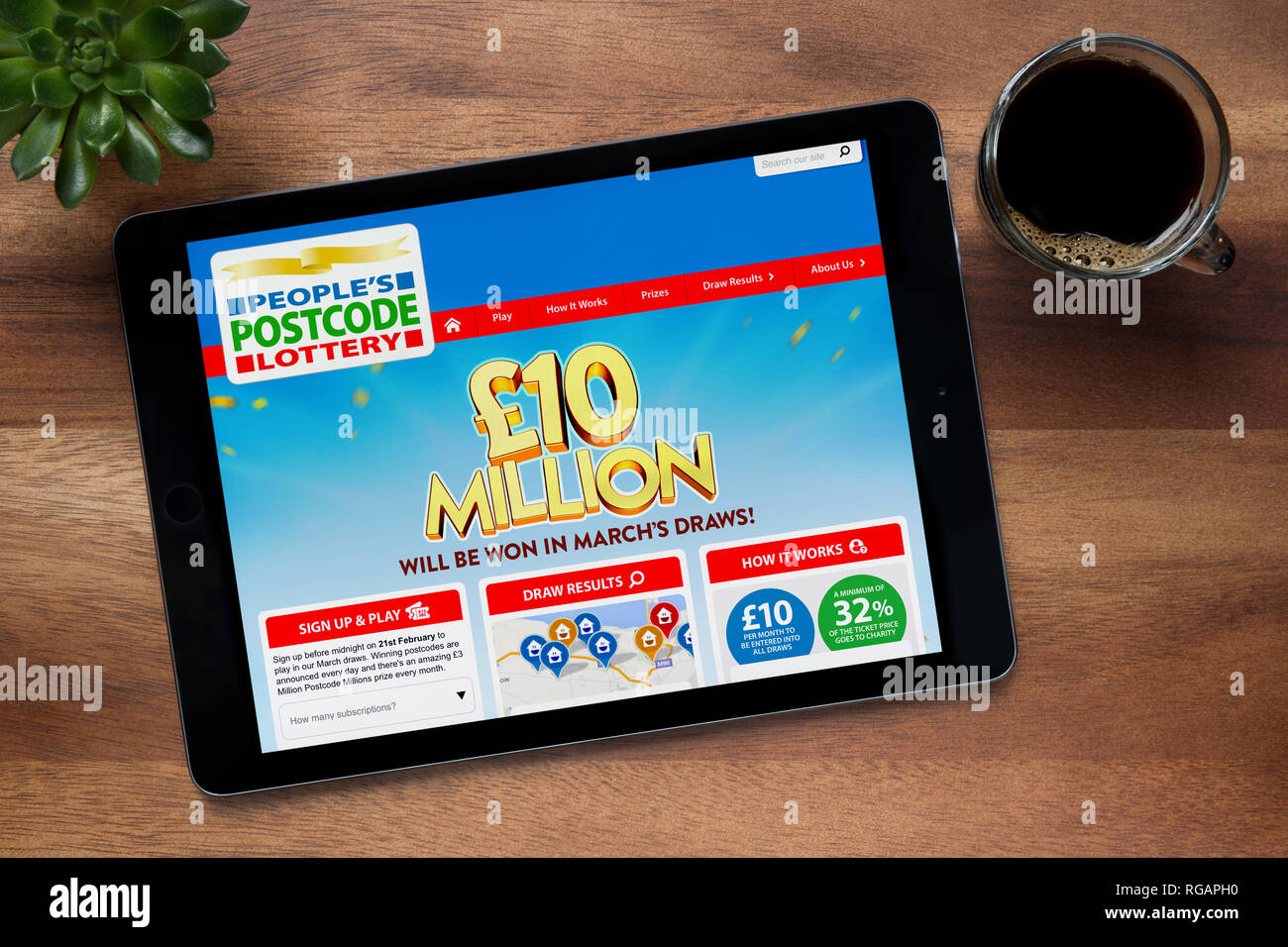 The website of People’s Postcode Lottery is seen on an iPad tablet, on a wooden table along with an espresso coffee and a house plant (Editorial only) Stock Photo