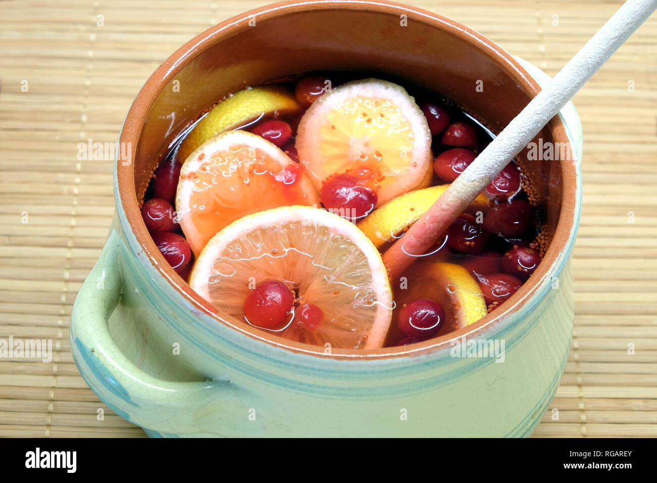 Non-alcoholic drink from cranberries and lemon in big round green pot with wooden spoon inside indoor on brown straw mat top view close up Stock Photo