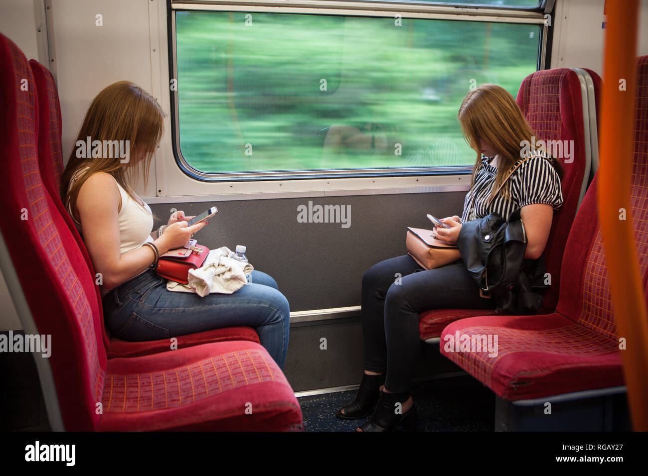 female twins in their 20's sitting oppossite on a train texting Stock Photo