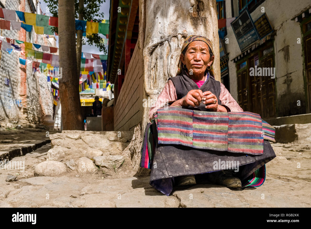 A local woman is sitting at a wall with prayer wheels and colorful prayer flags inside the walled town Stock Photo