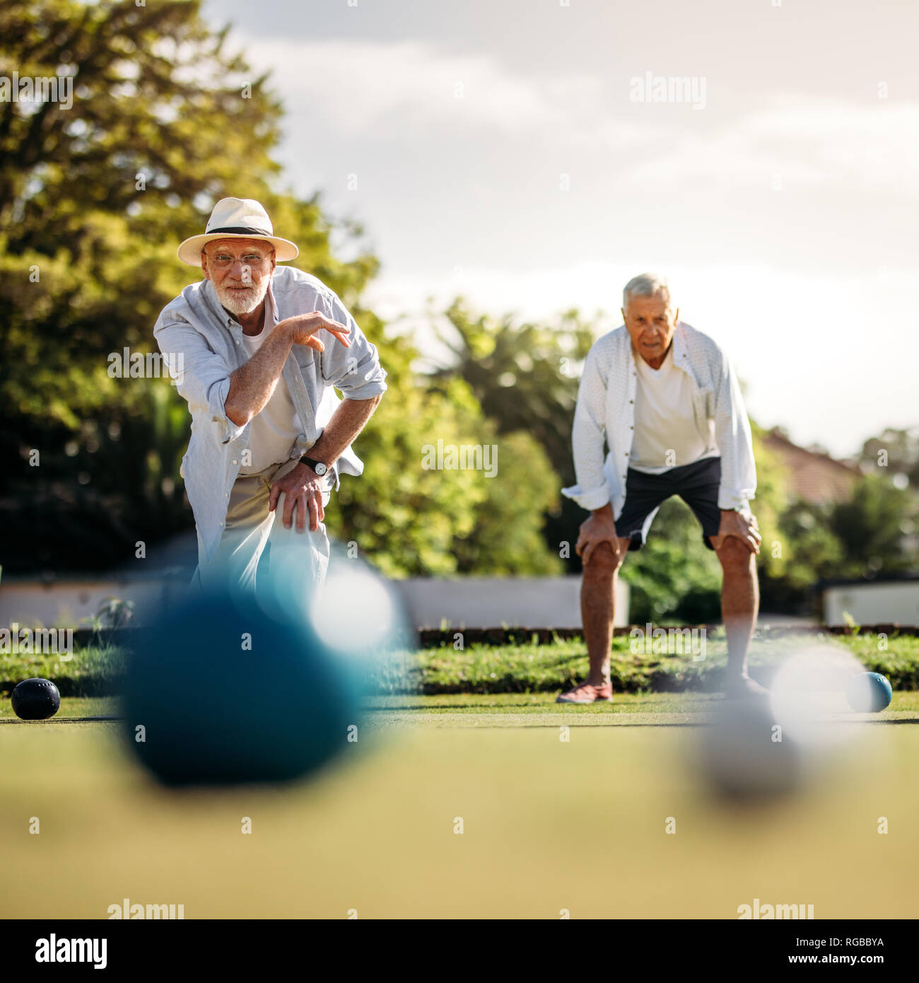 Elderly man playing boules in a playground with his playmate standing in the background. Old man in hat throwing a boules in a lawn with blurred boule Stock Photo