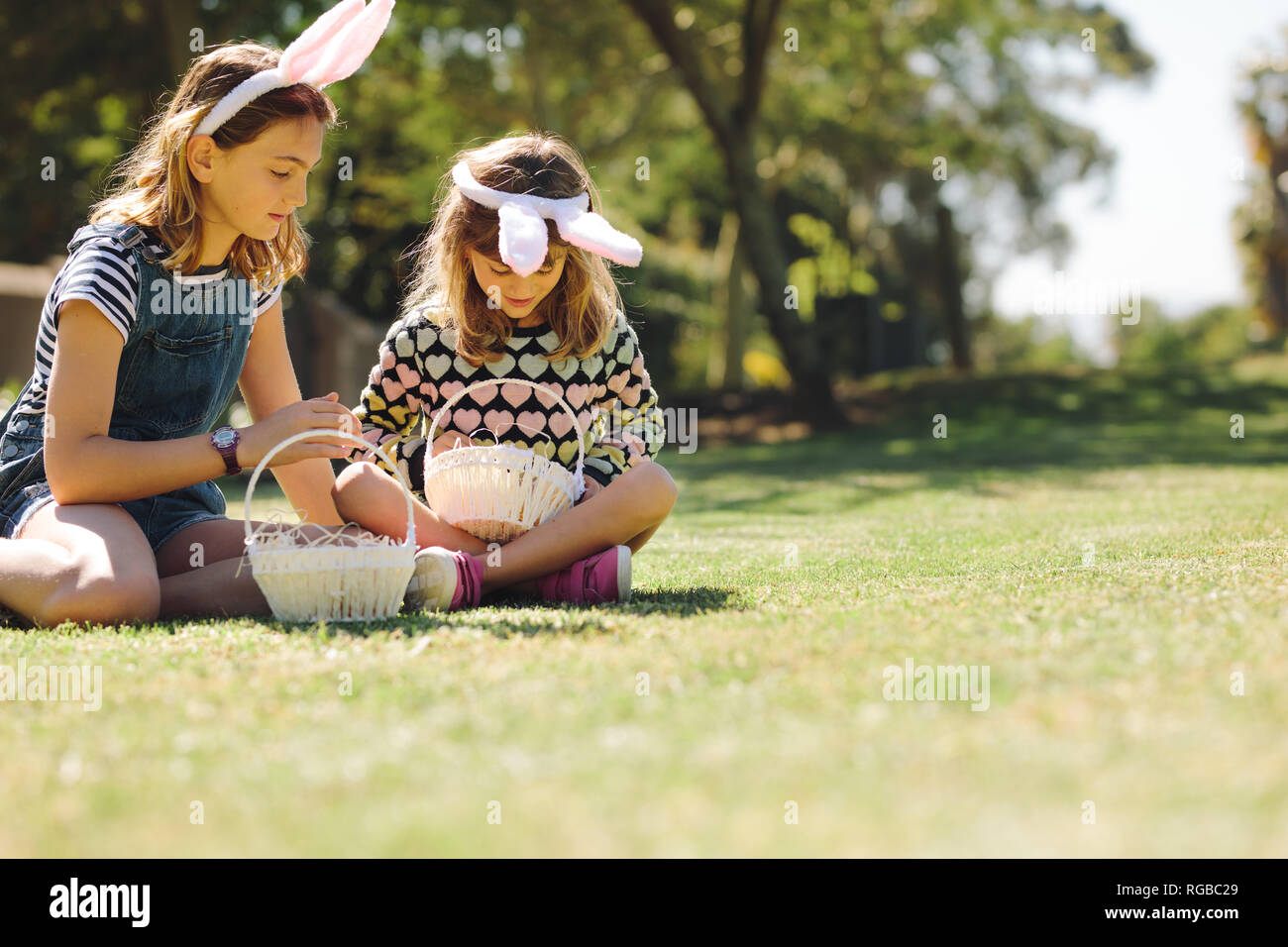 Two girls wearing rabbit ear headband playing in the backyard with baskets. Kids sitting on grass playing together on a sunny day. Stock Photo