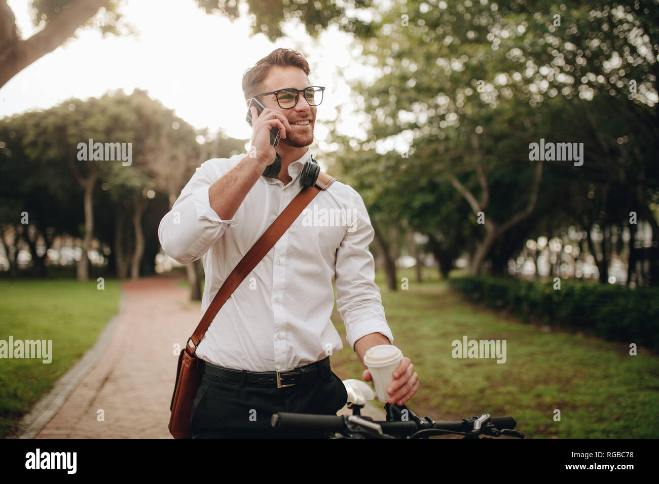 Smiling businessman talking over mobile phone standing on street with his bicycle. Man stopping by to make a call and drink coffee while commuting to Stock Photo