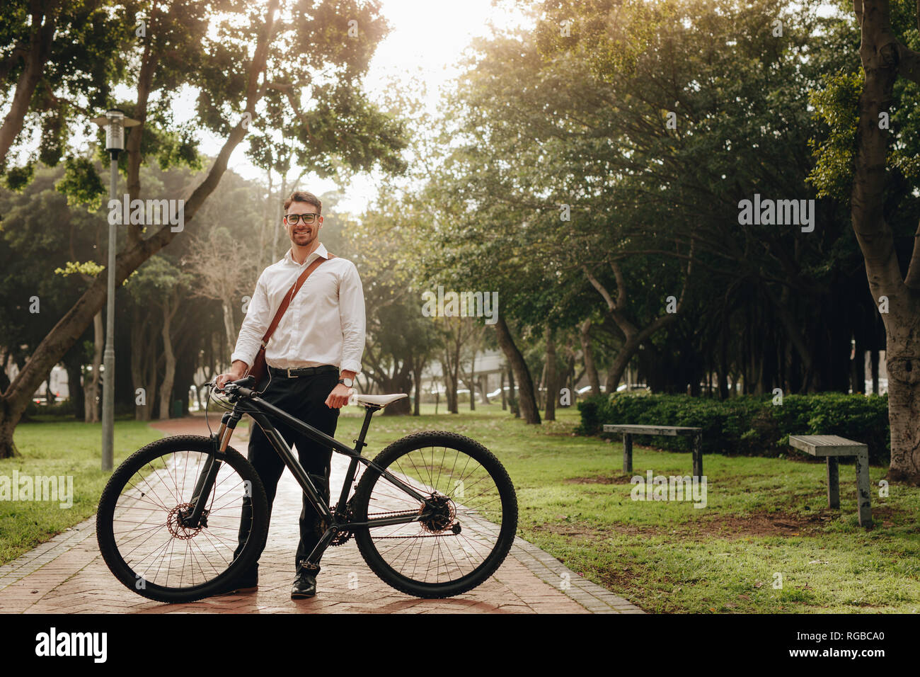 Businessman standing on the pathway of a park with his bicycle while going to office. Smiling man commuting to office on a bike standing in a park wit Stock Photo
