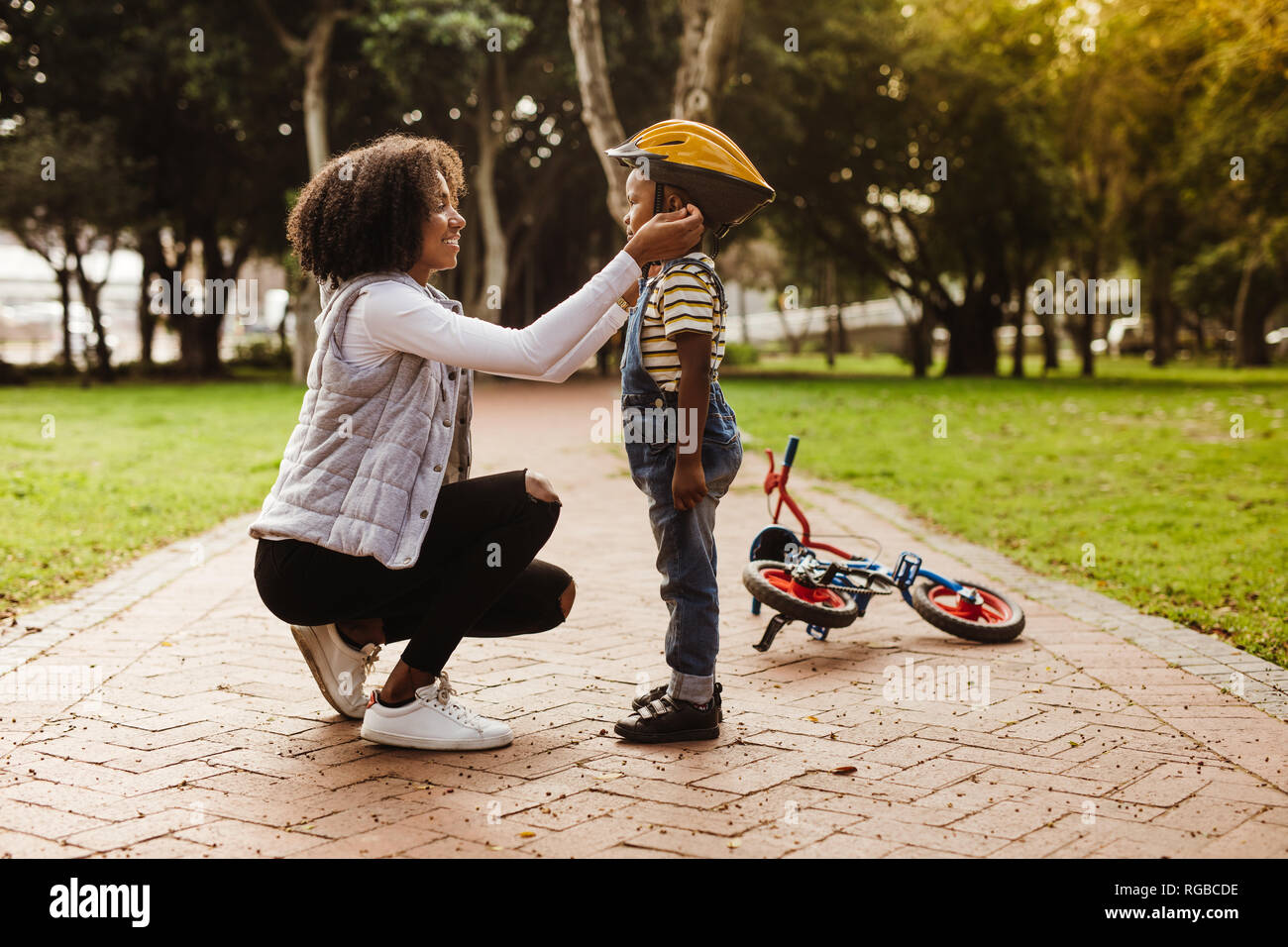 Young woman putting helmet on cute boy, with cycle lying at the back in park. Mother puts her son protective helmet for riding bike. Stock Photo
