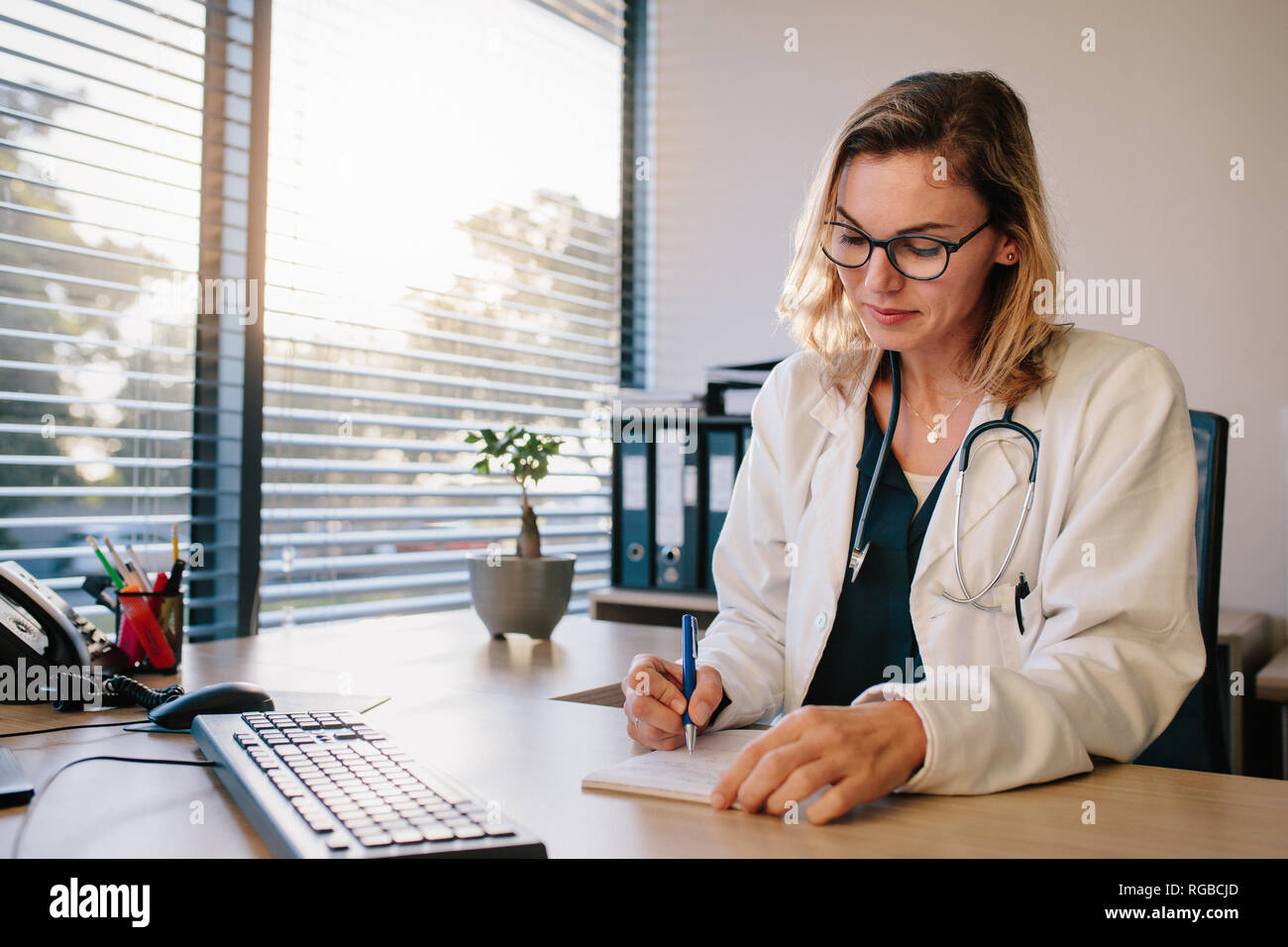 Female doctor sitting at the desk and writing prescription. Professional healthcare worker writing notes in a book. Stock Photo