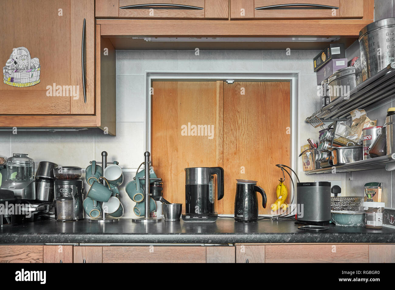 A Sonos speaker, cups, a kettle, a coffee grinder, a milk frother and other kitchen equipment on a cluttered work top in front of a oak hatch Stock Photo