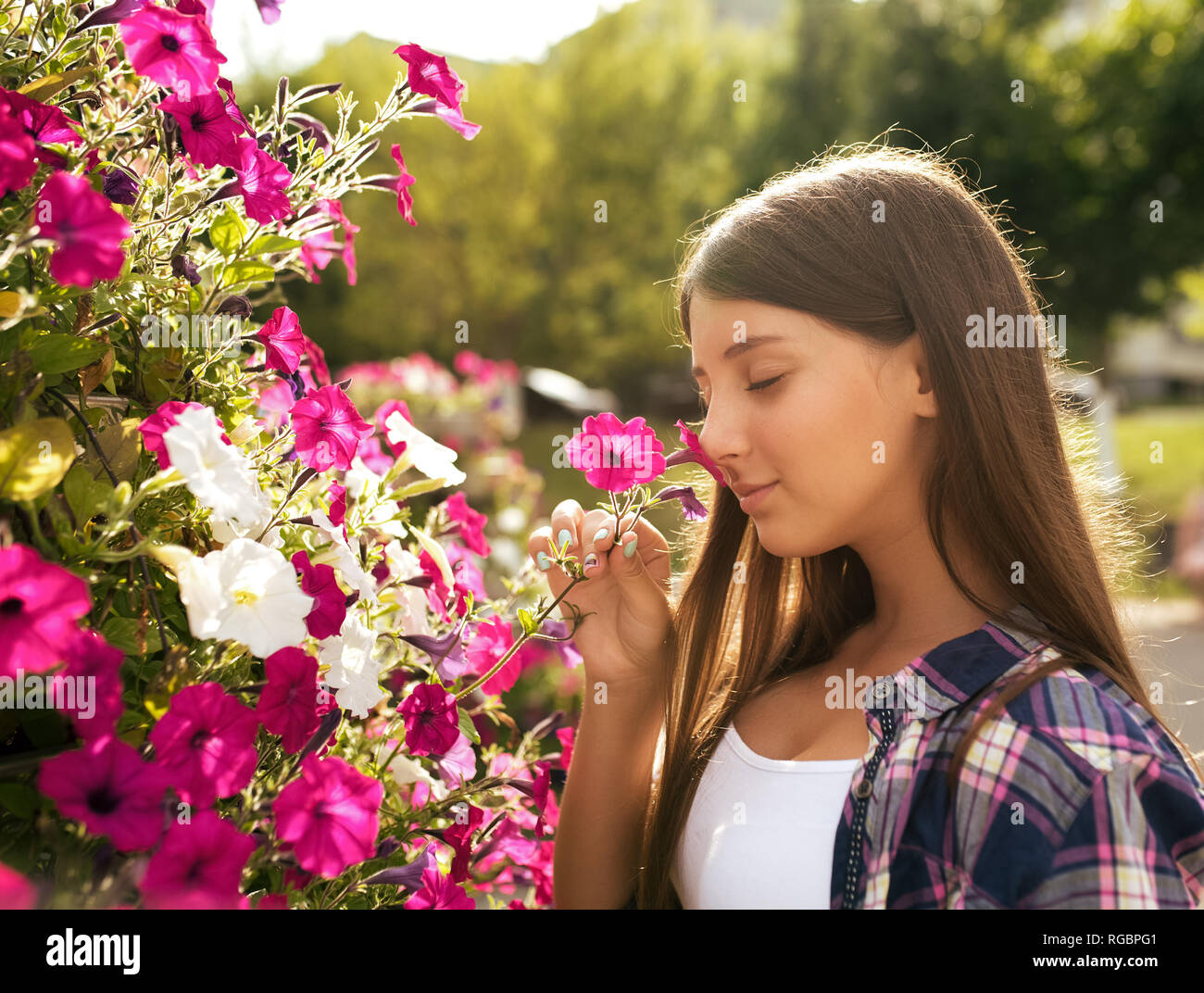A girl teenager 8-12 years old, stands in summer in a city park and smelling flowers in a flower bed. In summer, happy smiles. The concept of pleasant Stock Photo