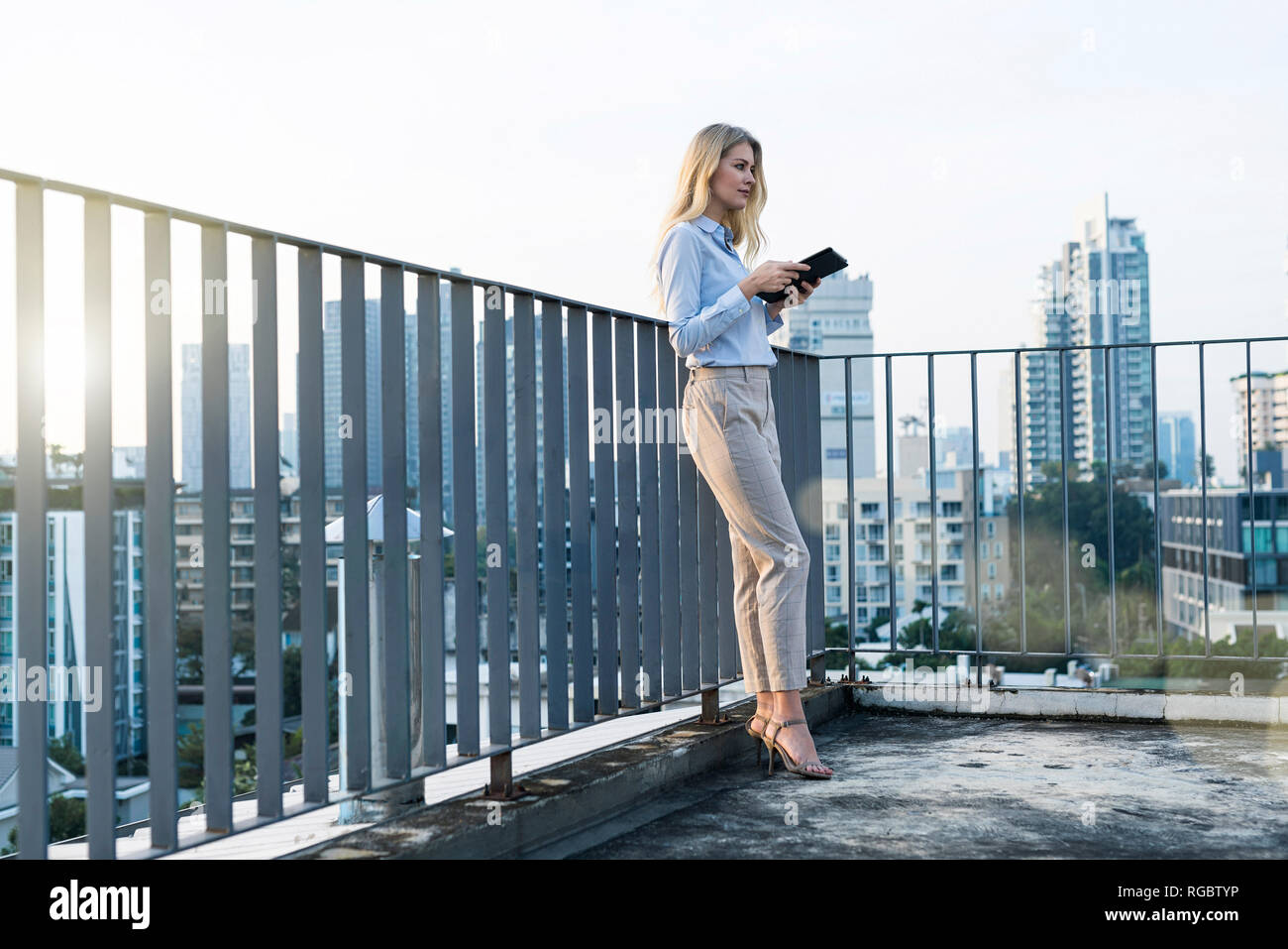 Blonde business woman leaning onto handrail holding tablet on city rooftop Stock Photo