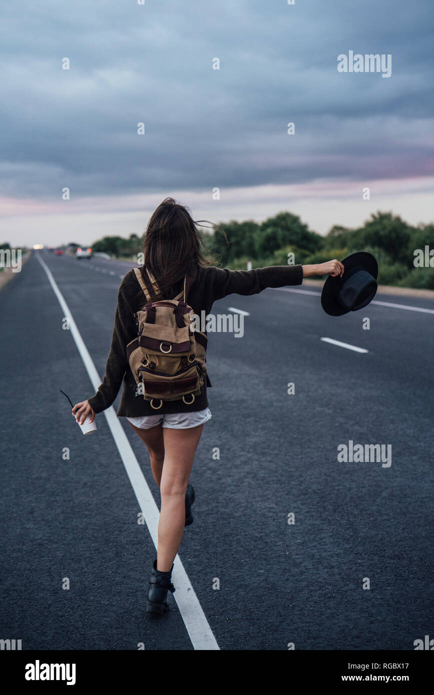 Back view of hitchhiking young woman with backpack and beverage standing at side line Stock Photo