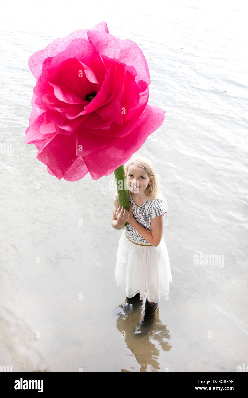 Portrait of smiling blond girl standing in a lake holding oversized pink artificial flower Stock Photo