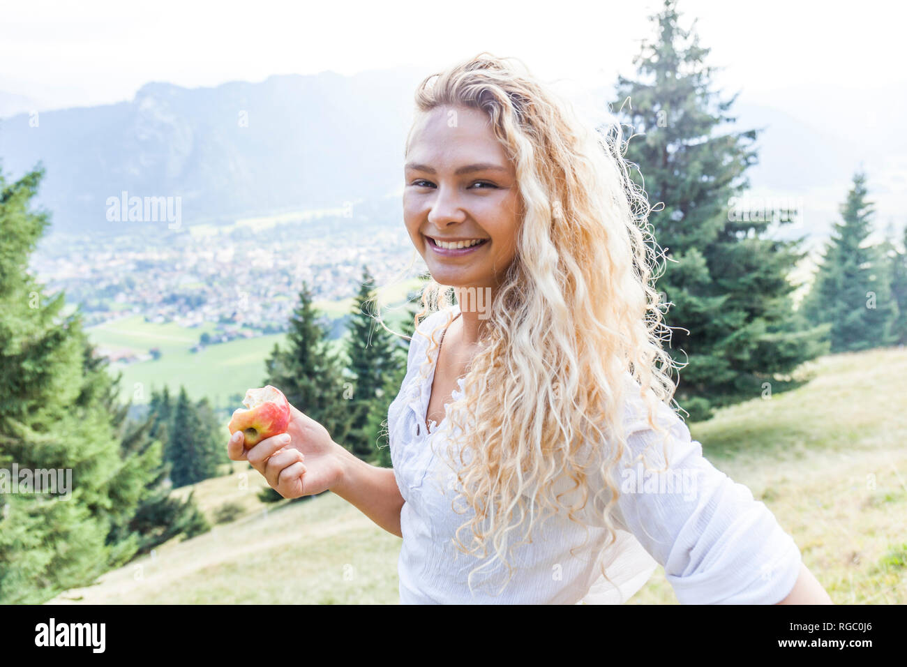 Germany, Bavaria, Oberammergau, portrait of smiling young woman eating an apple on mountain meadow Stock Photo