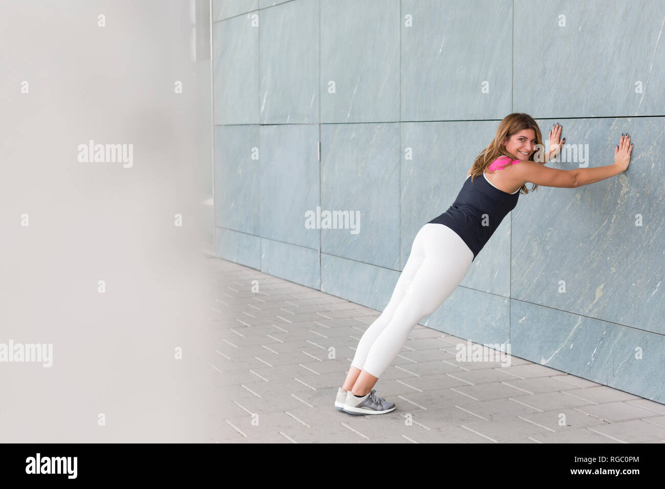 Smiling young woman leaning against wall doing stretching exercises hires stock photography and