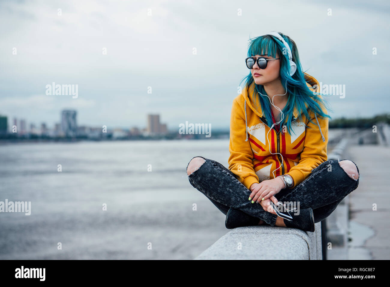 Young woman with dyed blue hair sitting on a wall listening music with headphones and smartphone Stock Photo