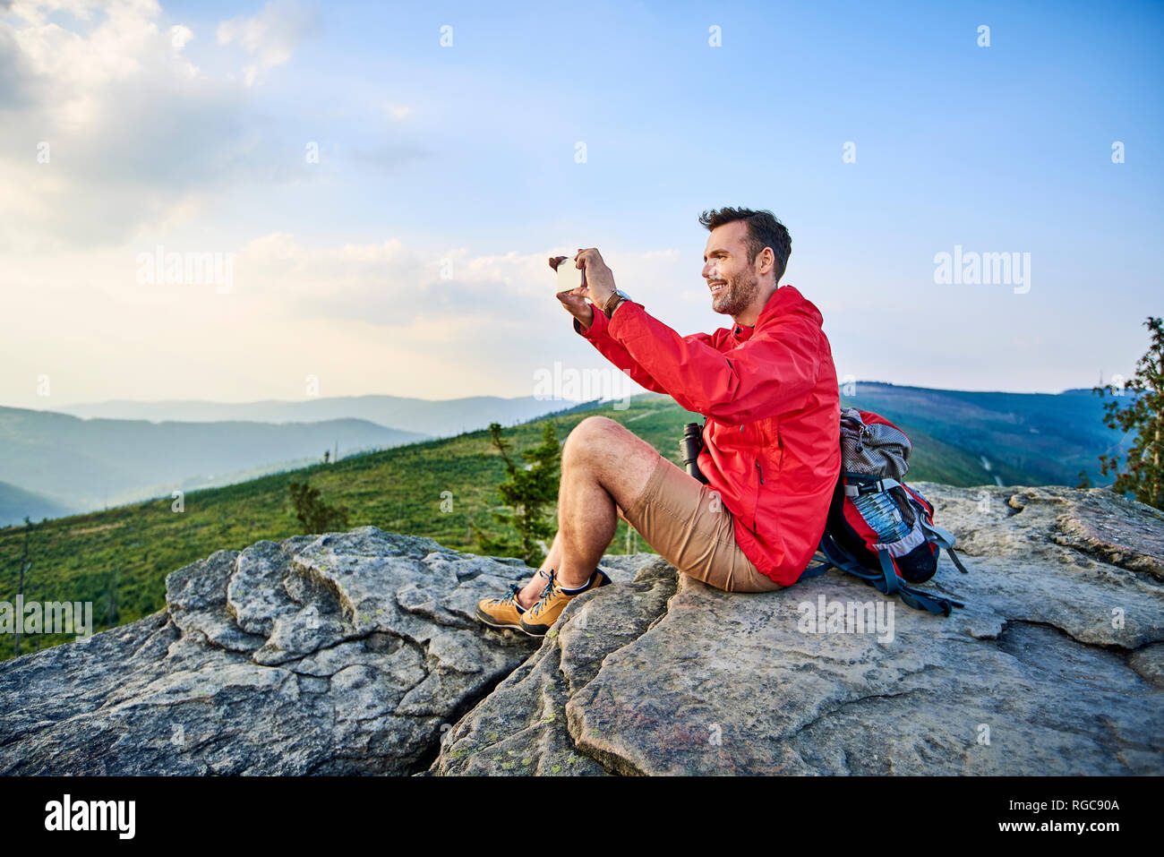 Man sitting on rock taking picture with his cell phone during hiking trip in the mountains Stock Photo