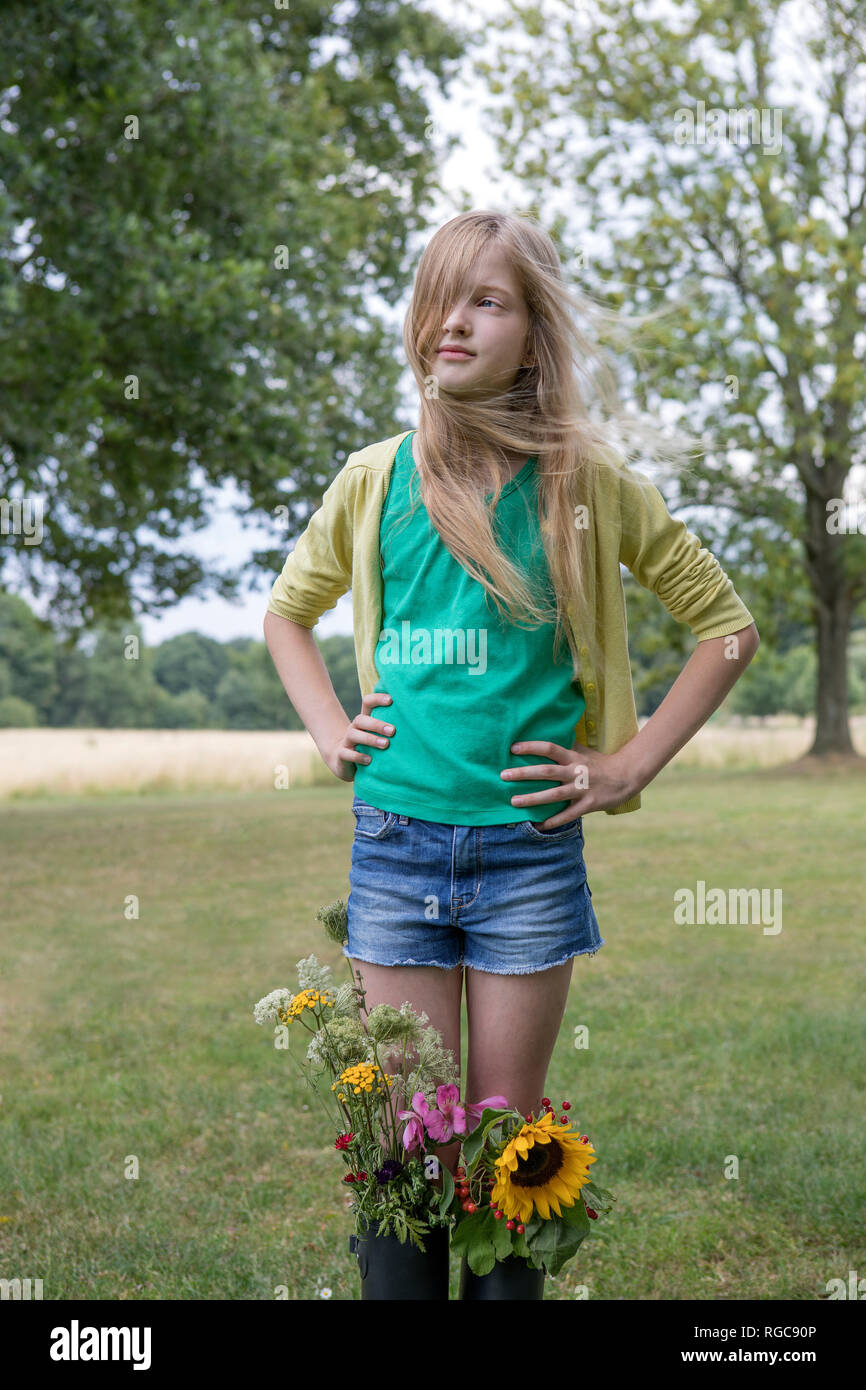 Portrait of blond girl standing on a meadow with bunches of flowers in her rubber boots Stock Photo