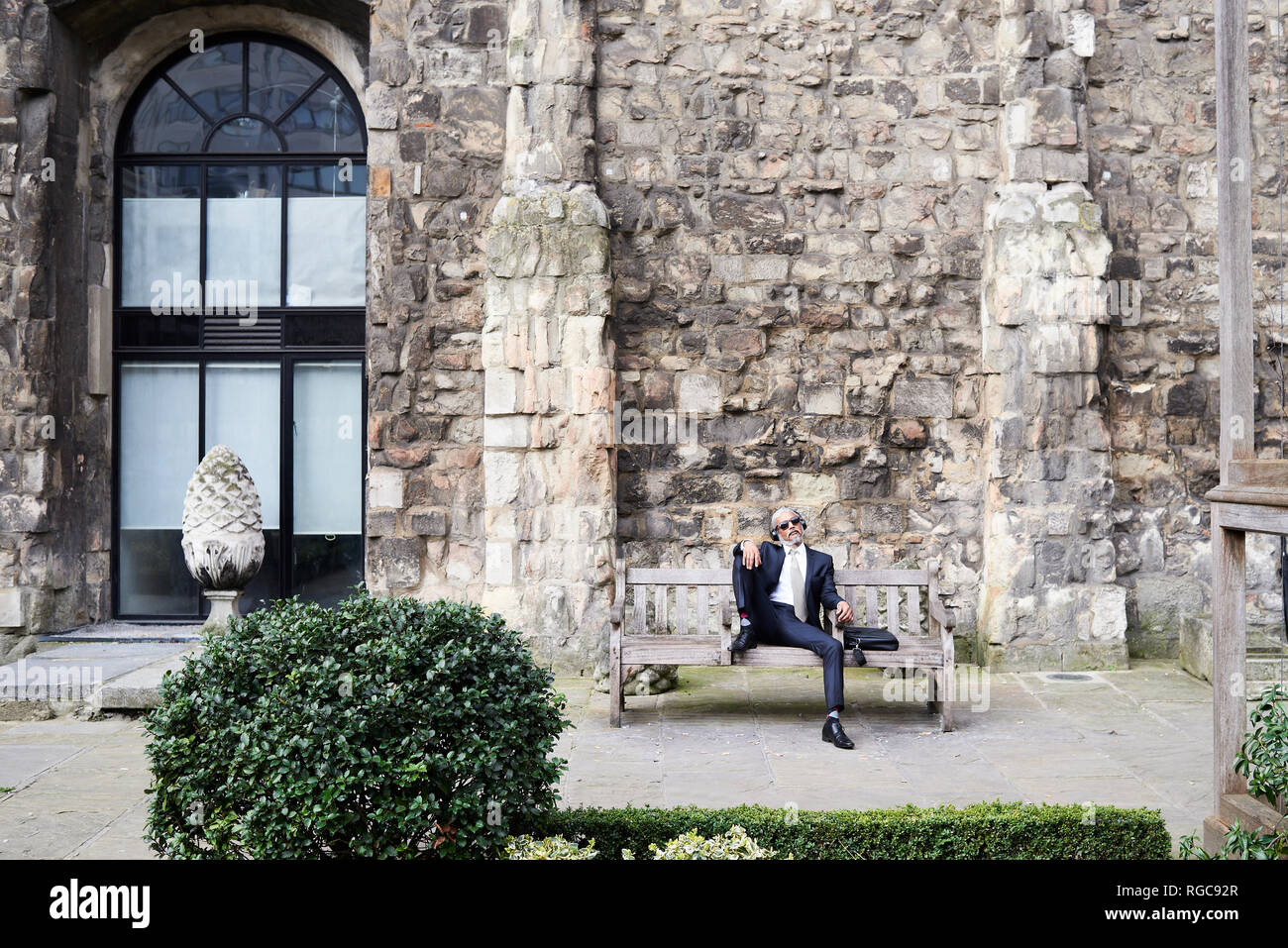 UK, London, senior businessman sitting on bench in a courtyard relaxing while liestening music with headphones Stock Photo