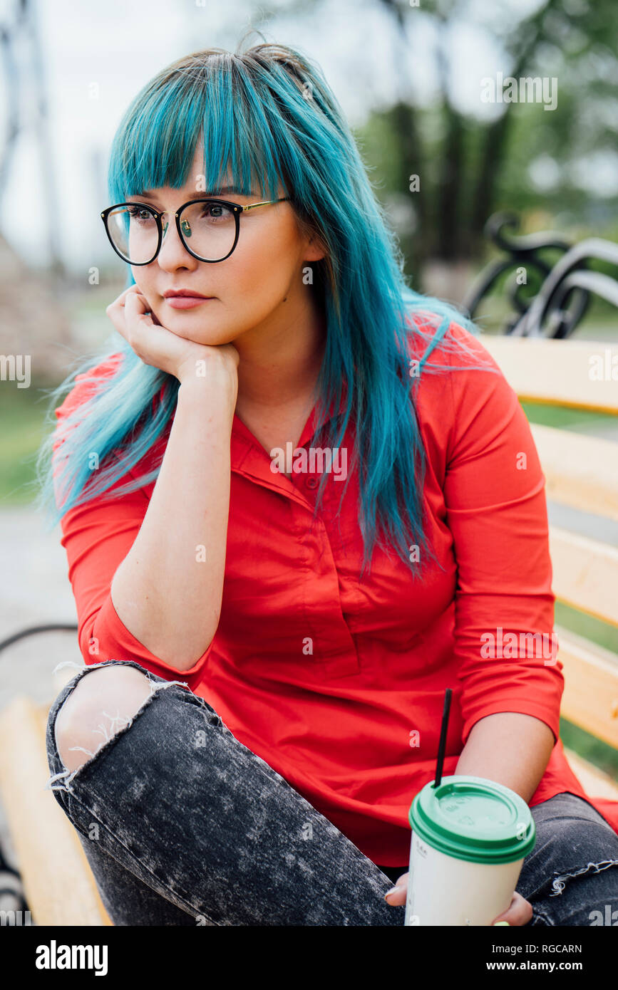 Portrait of young woman with dyed blue hair sitting on a bench with beverage Stock Photo